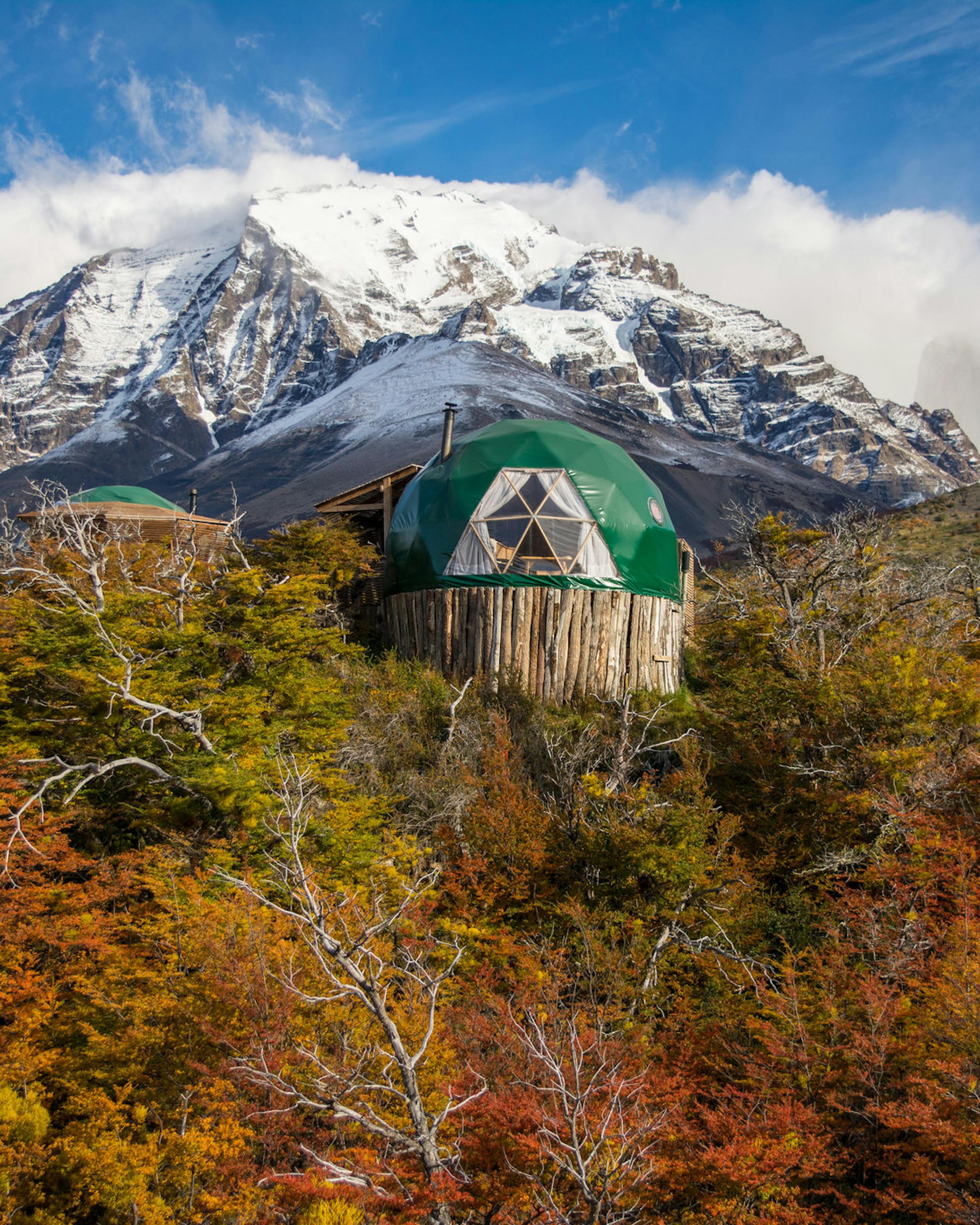 A green suite dome sits above autumn trees, backed by a snowcapped peak and clear Patagonian sky, with rugged peaks beyond.