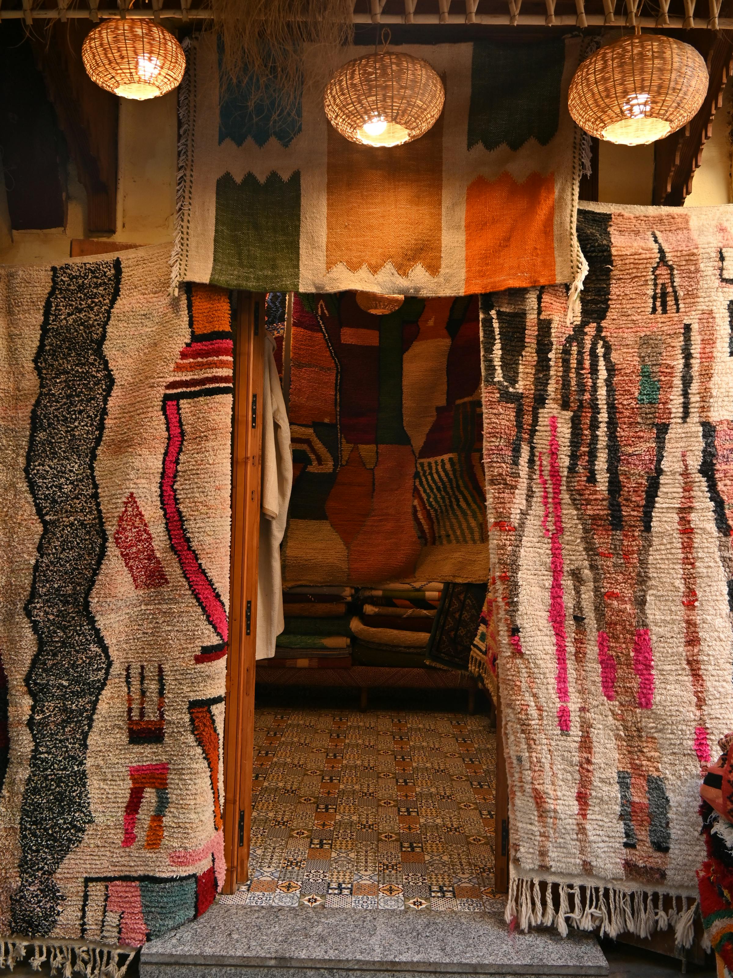 Colorful rugs hang at a medina shop entrance, framing a narrow doorway lined with patterned textiles and lanterns.