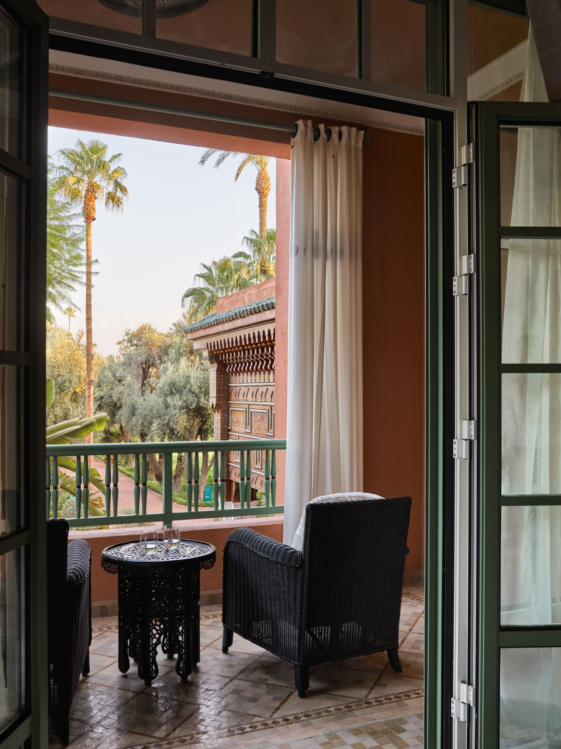 Open French doors lead to a private balcony at La Mamounia, looking over palms and manicured gardens in daylight.