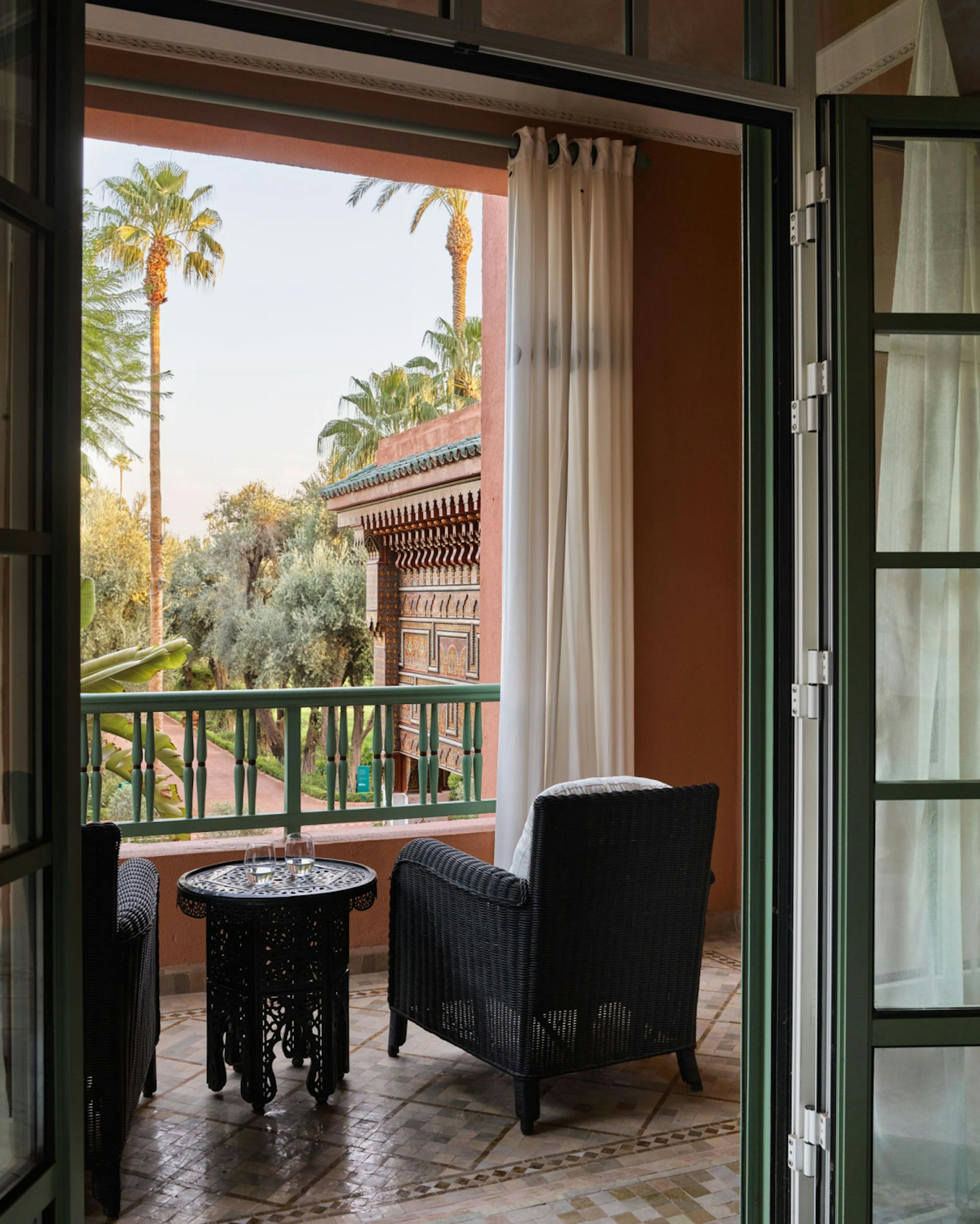 Open French doors lead to a private balcony at La Mamounia, looking over palms and manicured gardens in daylight.