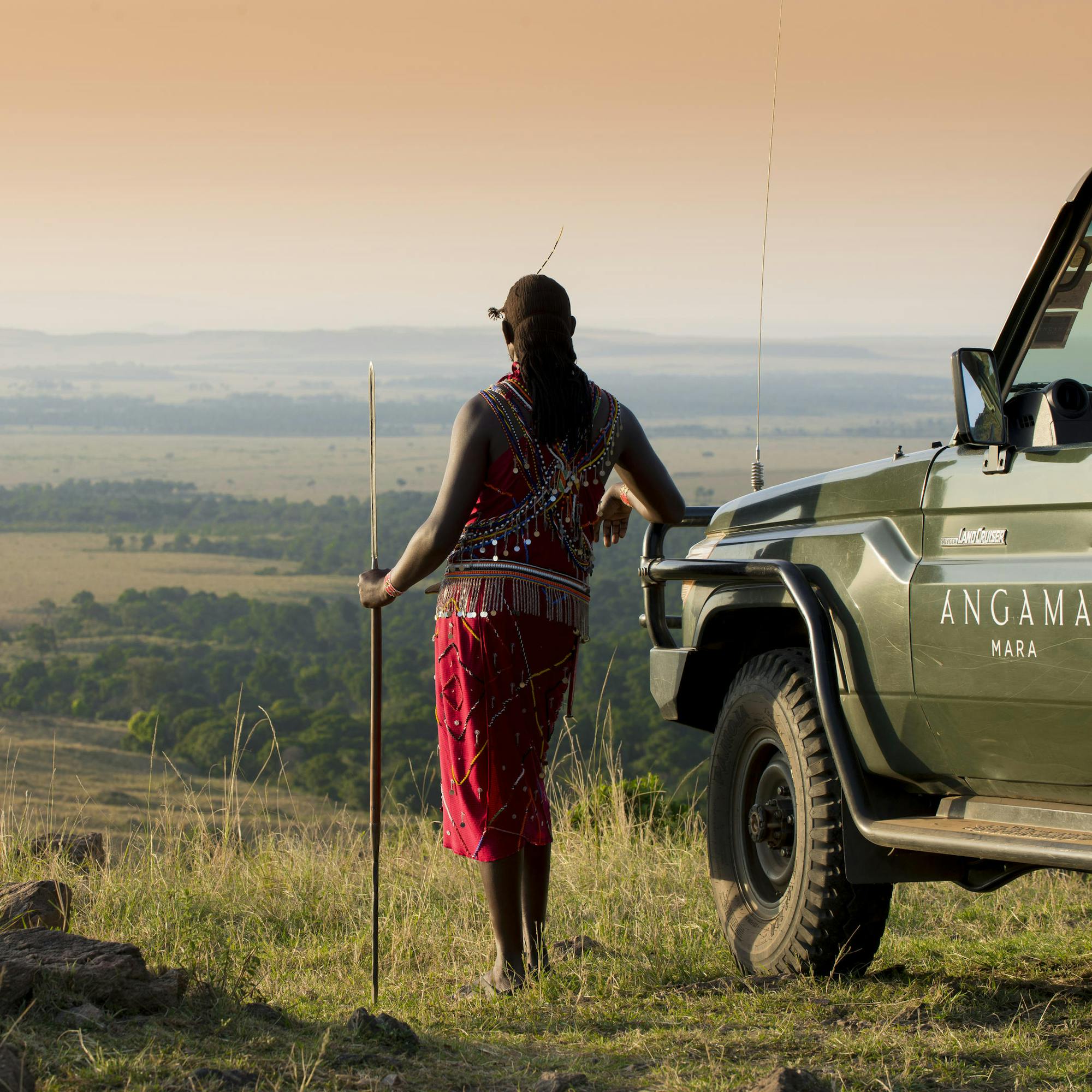 A Maasai guide in a red shuka walks toward an open safari vehicle at a viewpoint above the Mara in warm golden light.