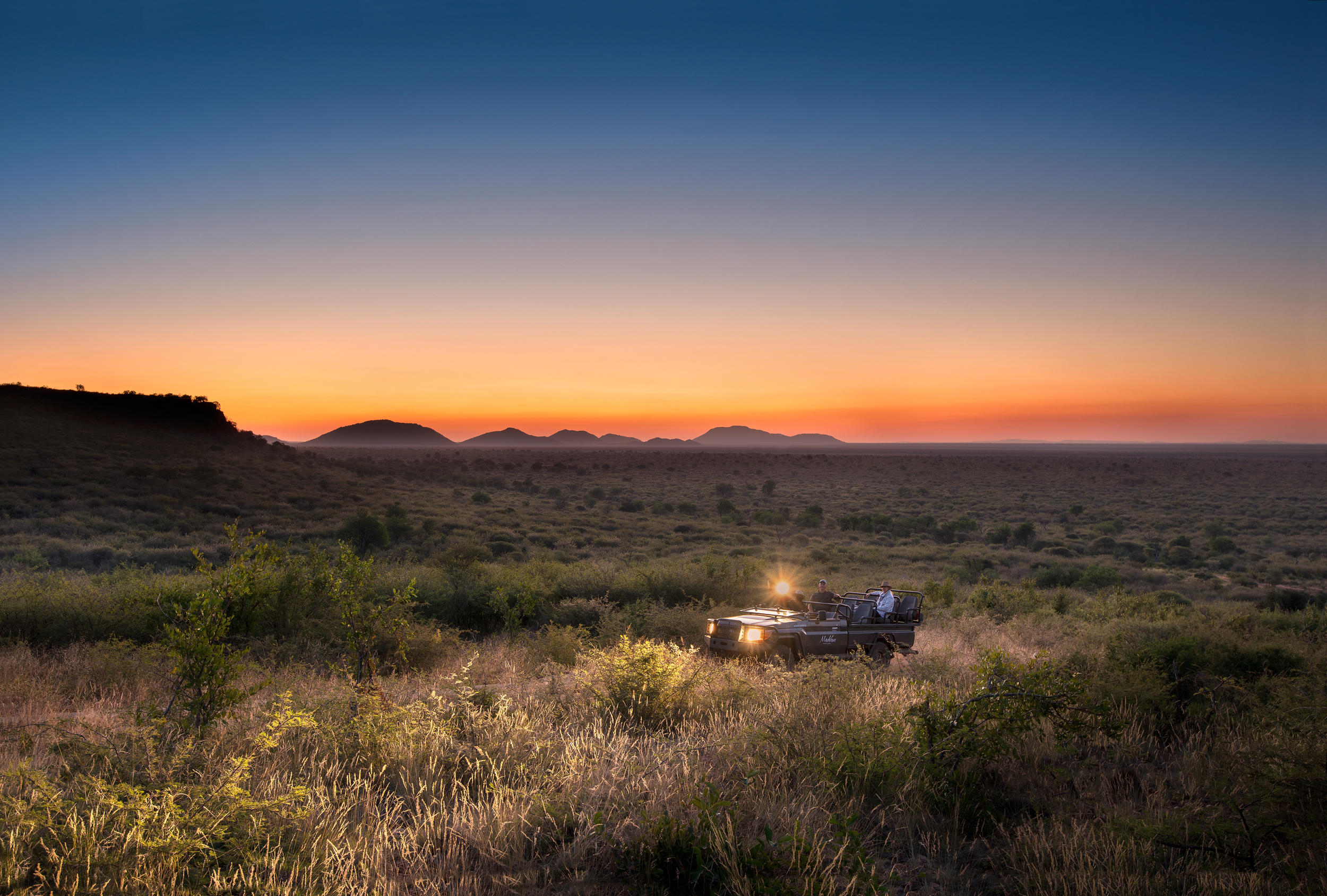 A safari vehicle drives through tall grass at sunset, headlights glowing as the horizon fades to orange.