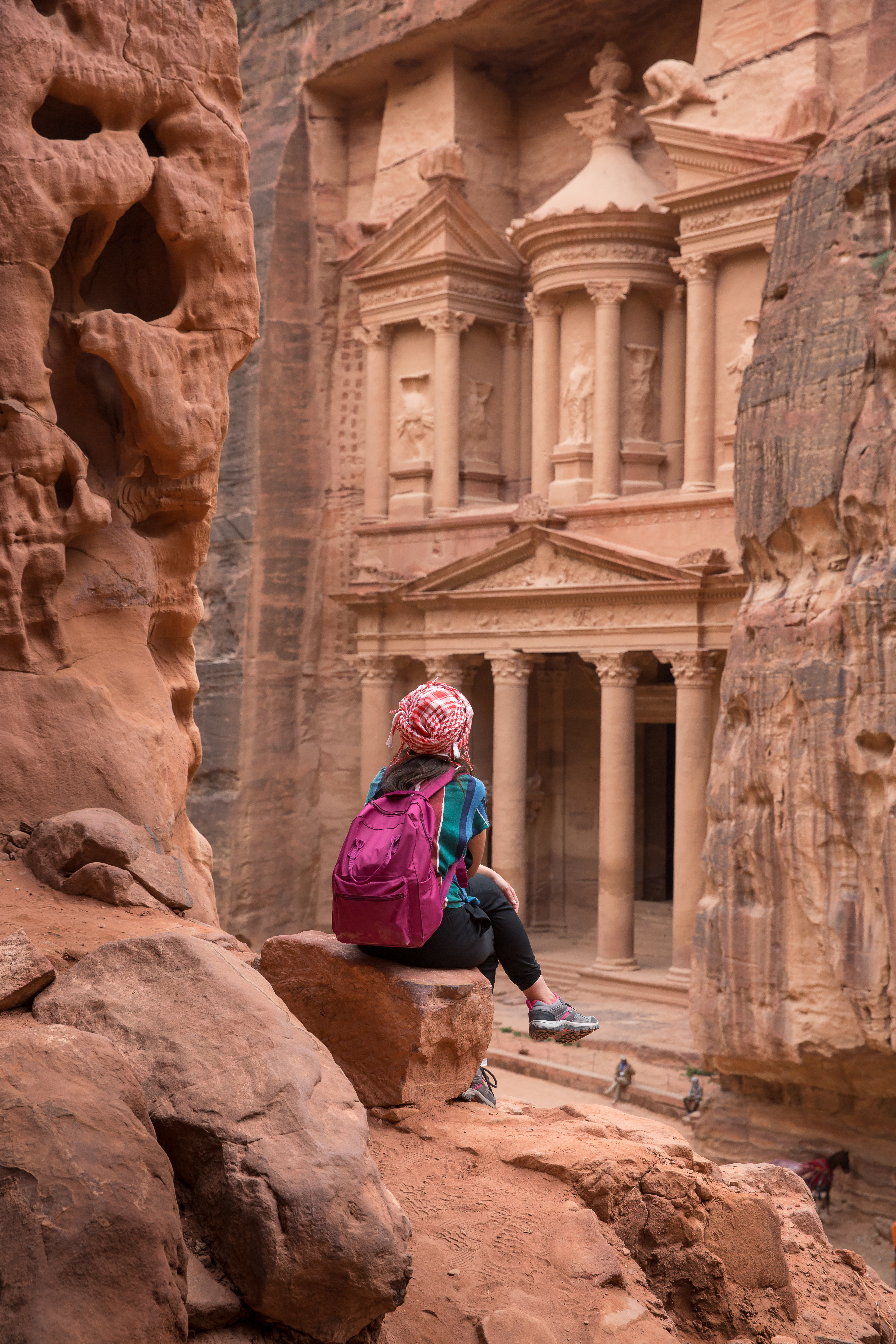 A traveler with a backpack sits on a rock ledge, looking out at Petra’s Treasury framed by towering canyon walls.