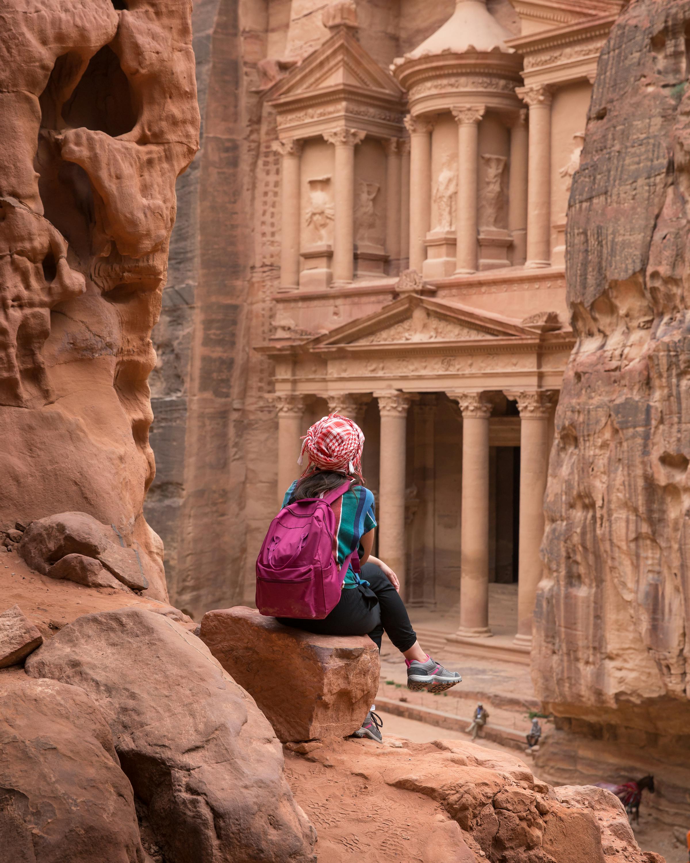 A traveler with a backpack sits on a rock ledge, looking out at Petra’s Treasury framed by towering canyon walls.