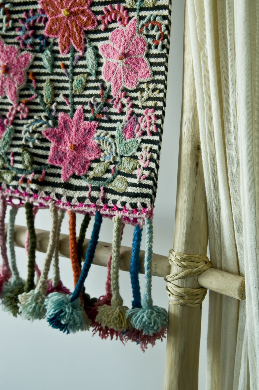 Colorful woven textile hangs beside a window, bold floral patterns and fringe highlighted in soft indoor light.