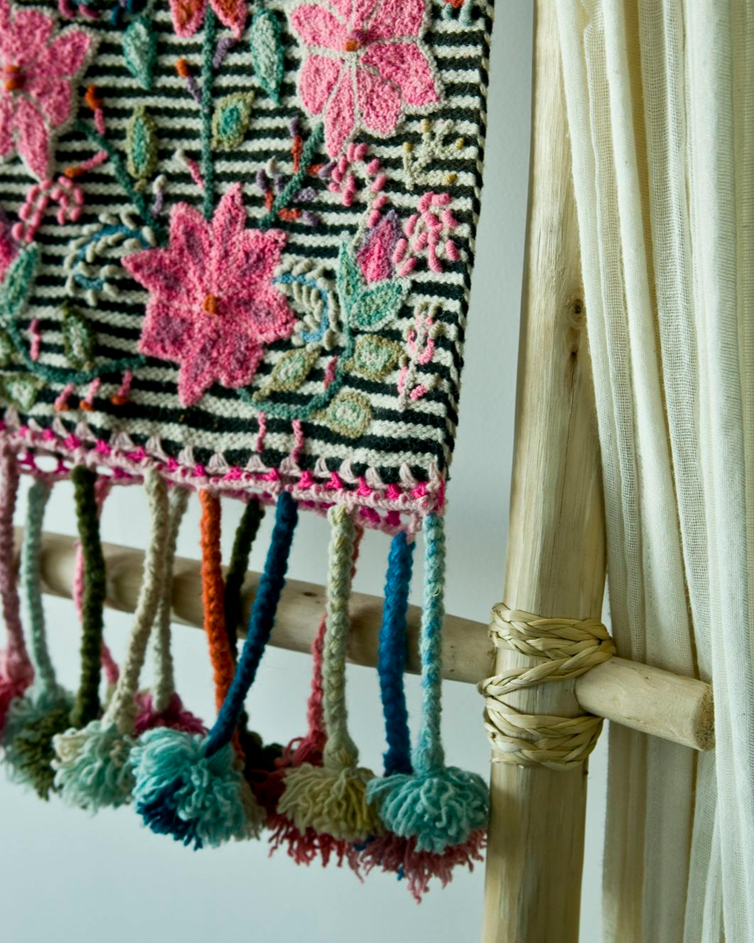 Colorful woven textile hangs beside a window, bold floral patterns and fringe highlighted in soft indoor light.