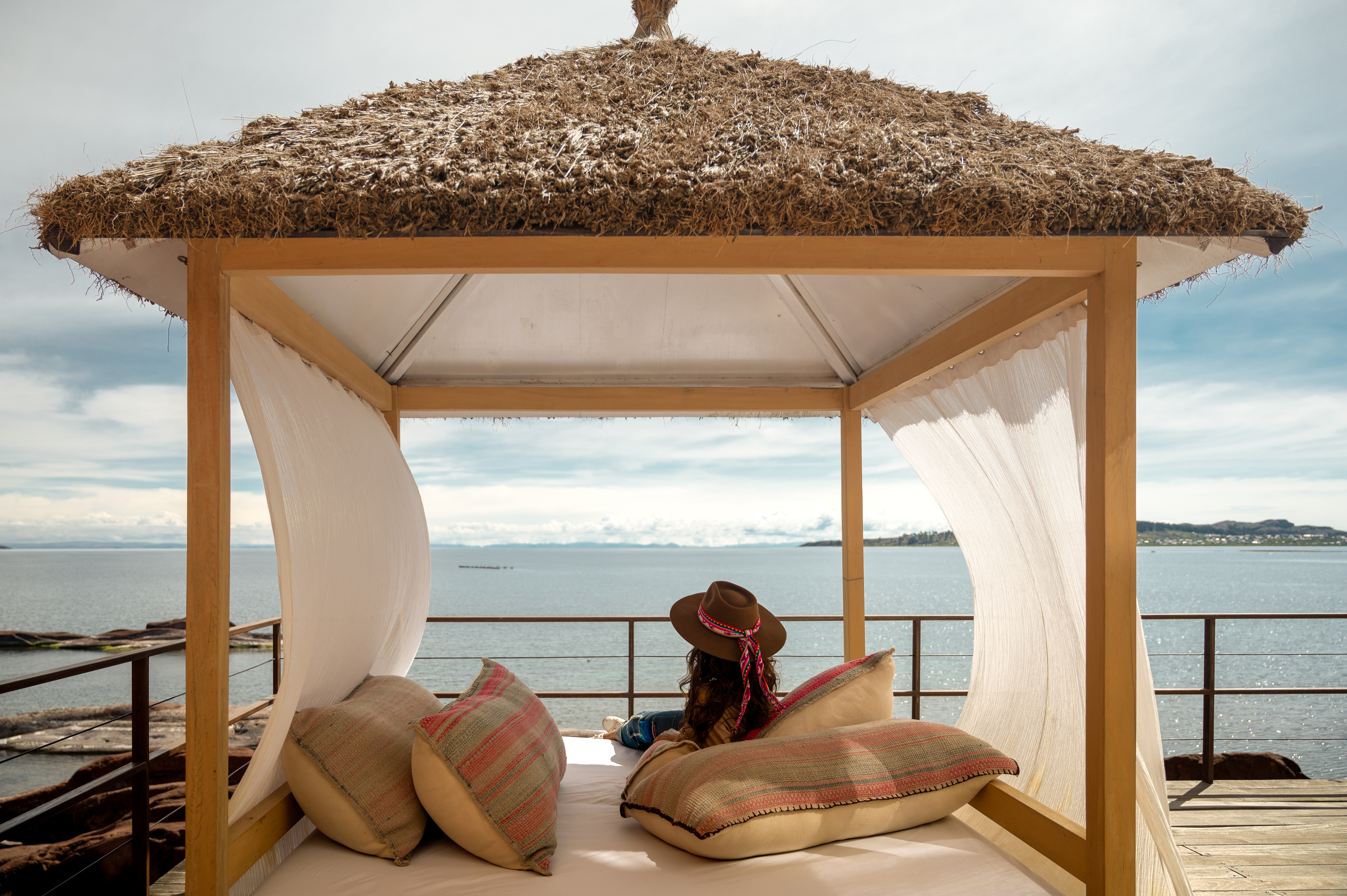 Traveler reclines on a cushioned daybed beneath a thatched cabana, facing Lake Titicaca and mountains at sunset.