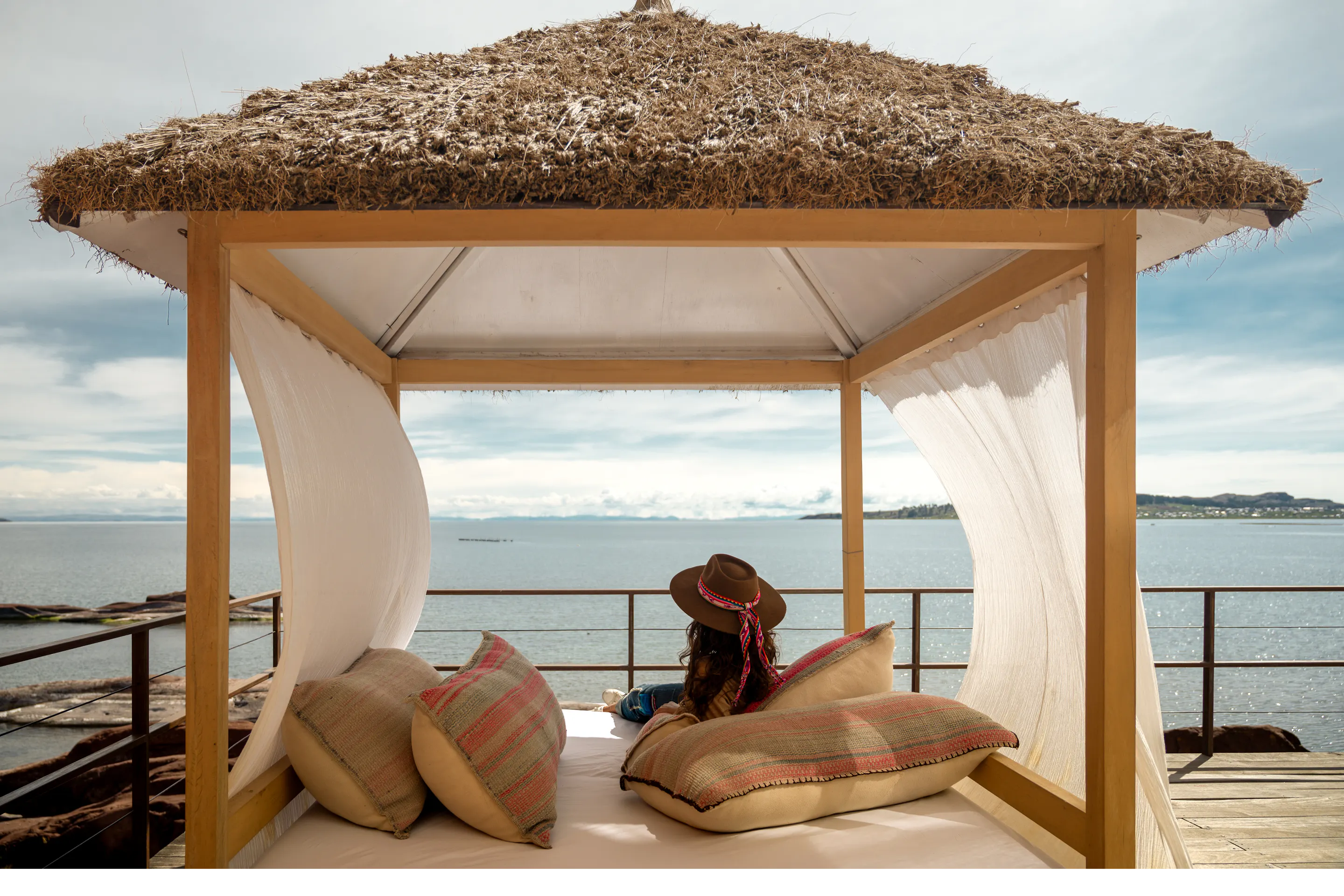 Traveler reclines on a cushioned daybed beneath a thatched cabana, facing Lake Titicaca and mountains at sunset.