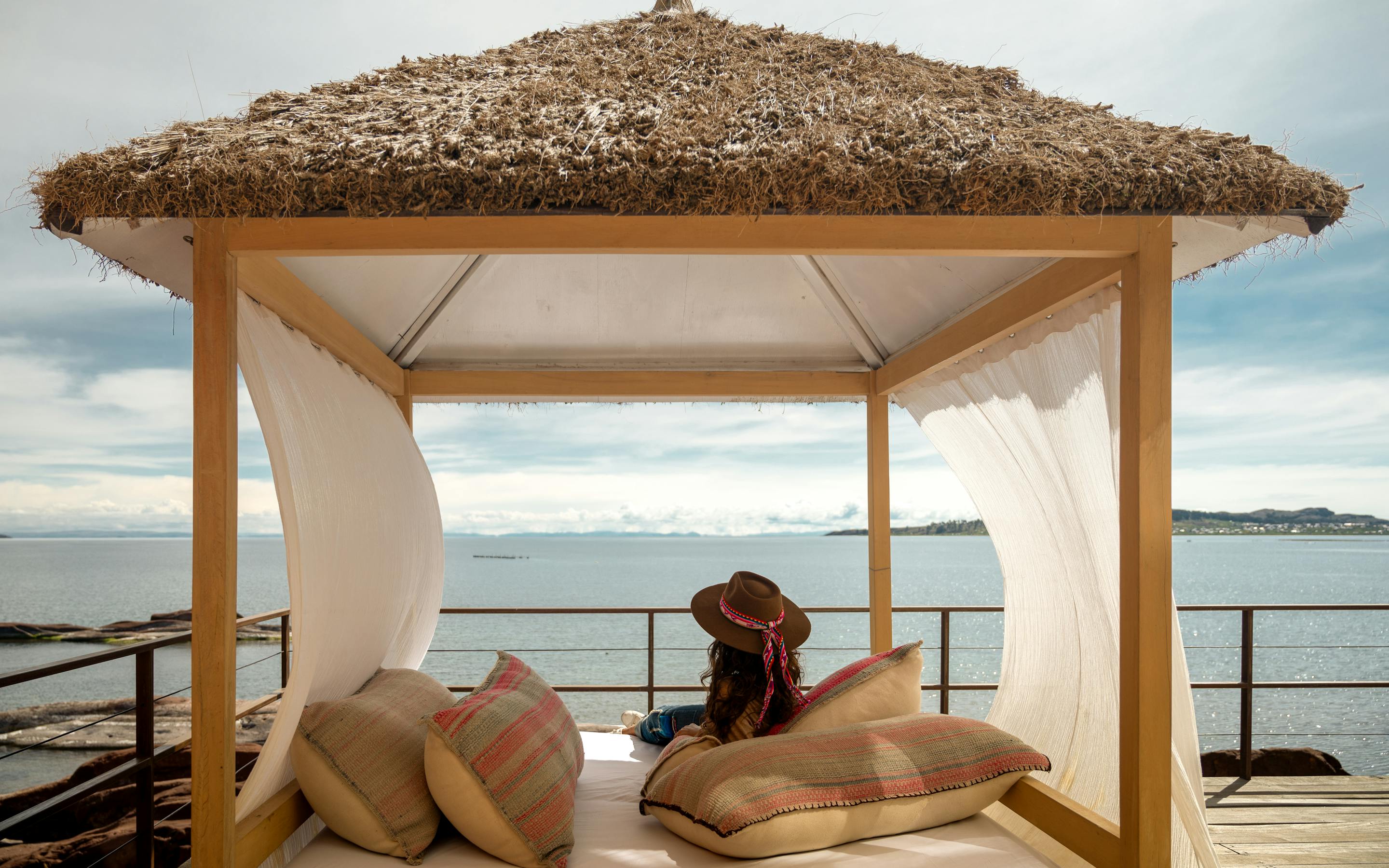 Traveler reclines on a cushioned daybed beneath a thatched cabana, facing Lake Titicaca and mountains at sunset.