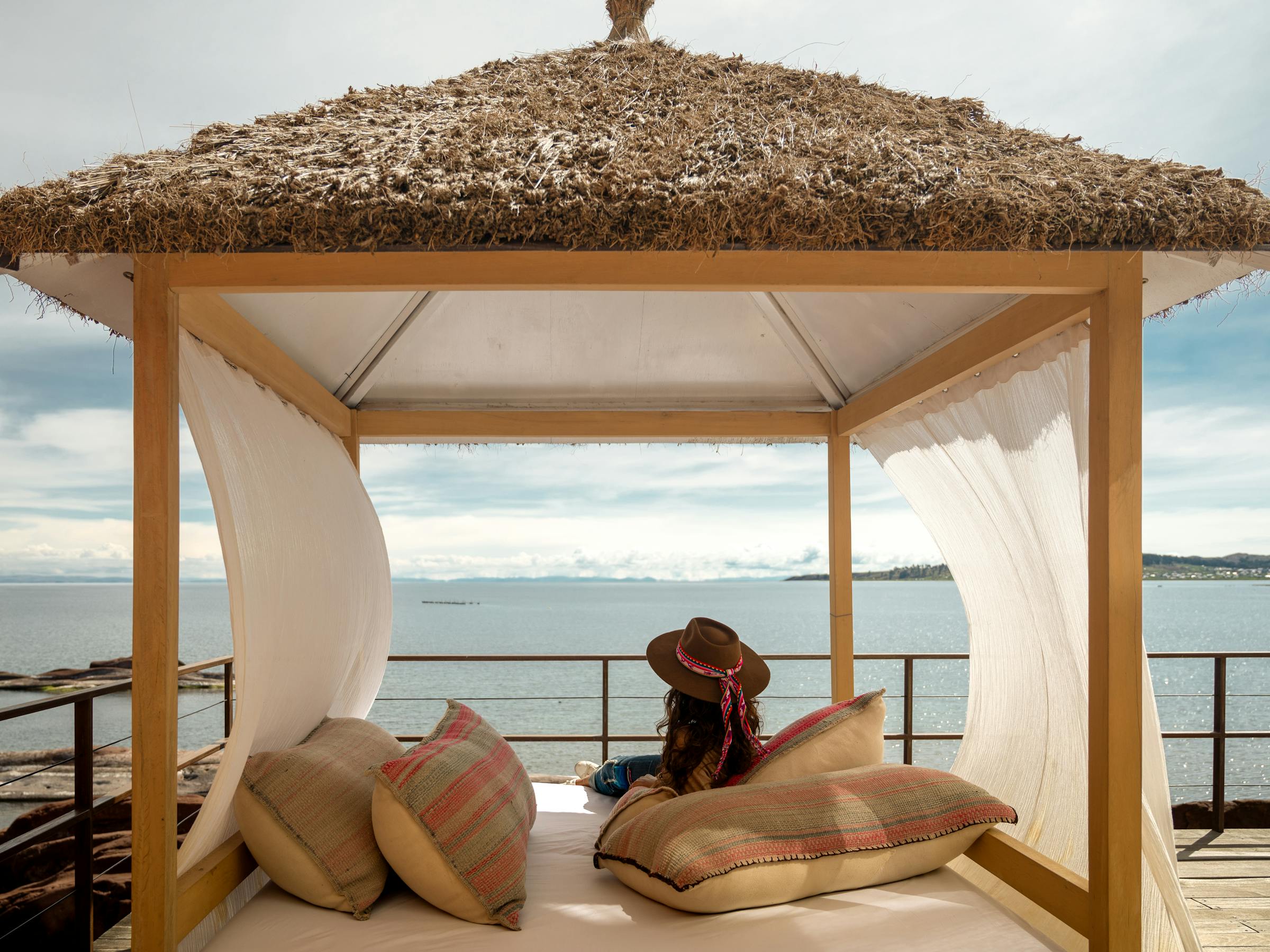 Traveler reclines on a cushioned daybed beneath a thatched cabana, facing Lake Titicaca and mountains at sunset.