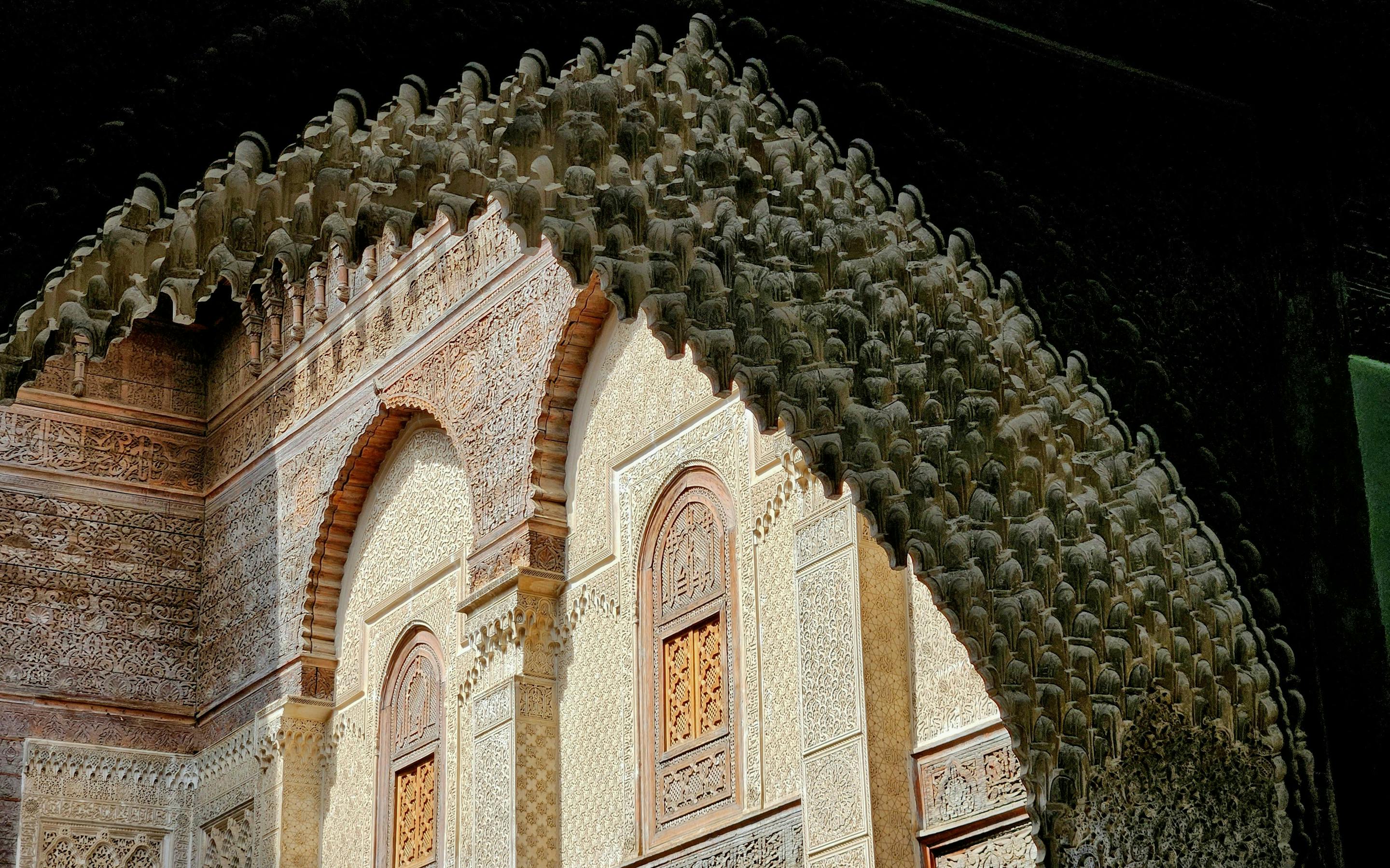Sunlight falls across a horseshoe arch with carved plasterwork and wooden lattice windows beneath a deep overhang.