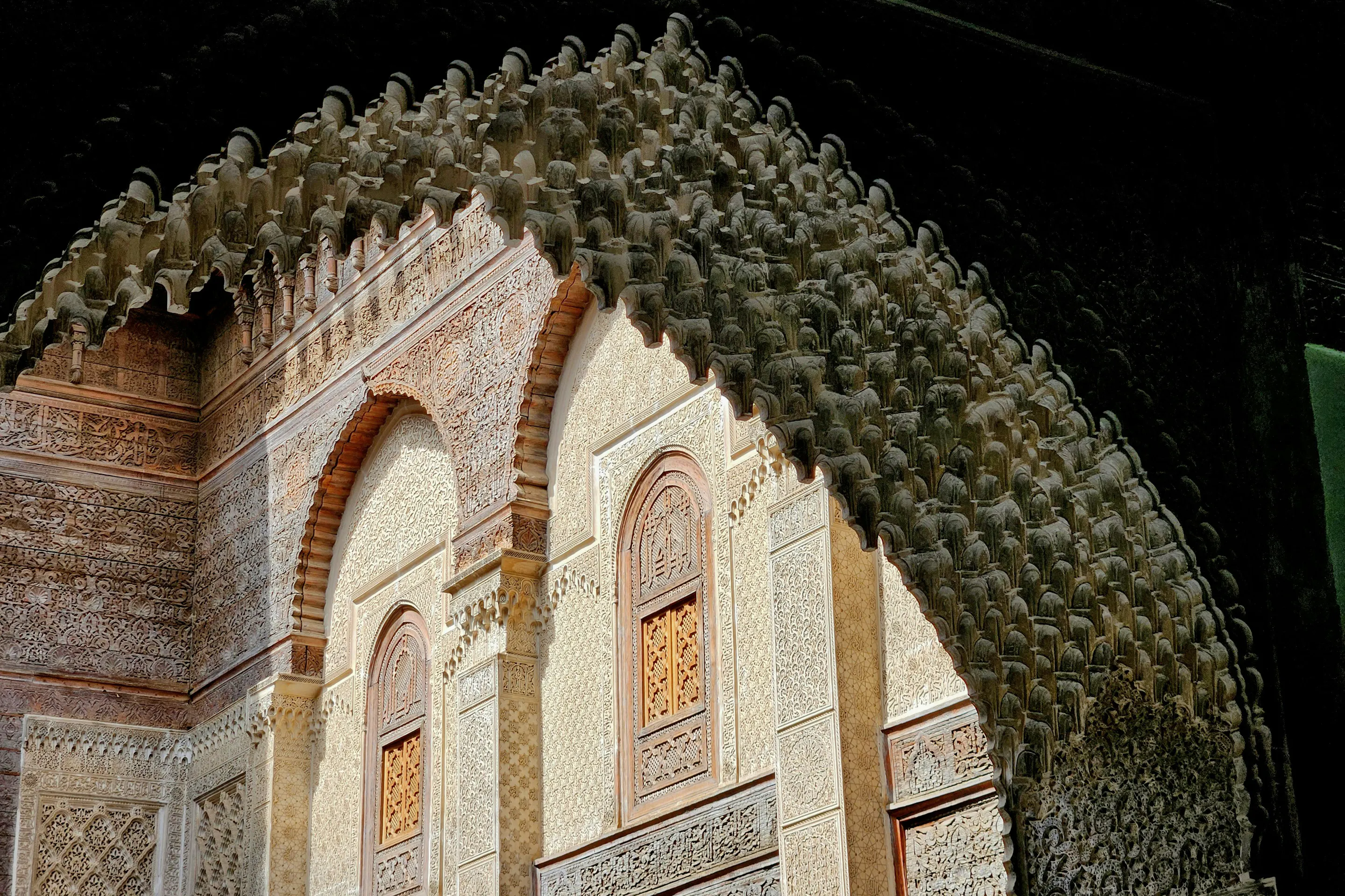 Sunlight falls across a horseshoe arch with carved plasterwork and wooden lattice windows beneath a deep overhang.