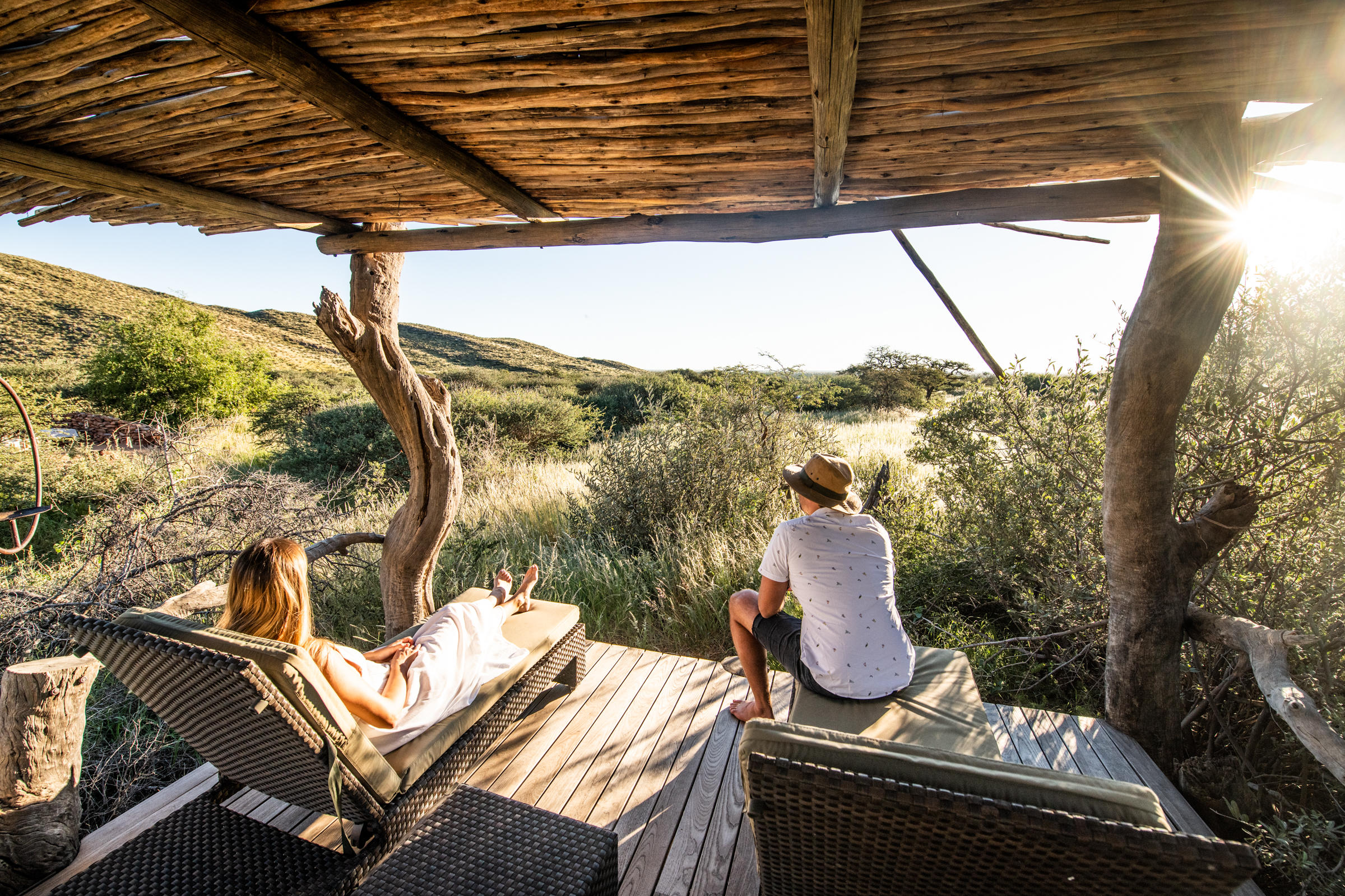 Two guests relax under a thatched shelter, enjoying sundowners as the Kalahari turns gold at sunset, in warm evening light.