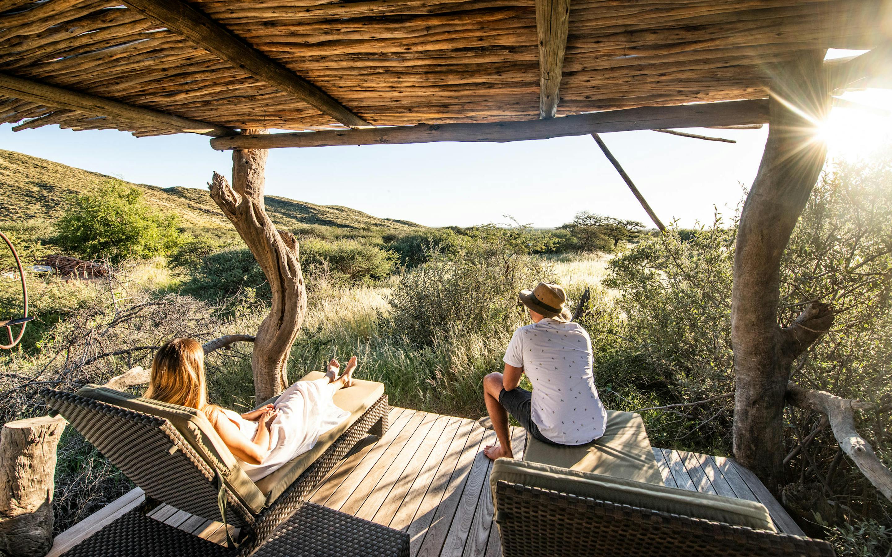 Two guests relax under a thatched shelter, enjoying sundowners as the Kalahari turns gold at sunset, in warm evening light.