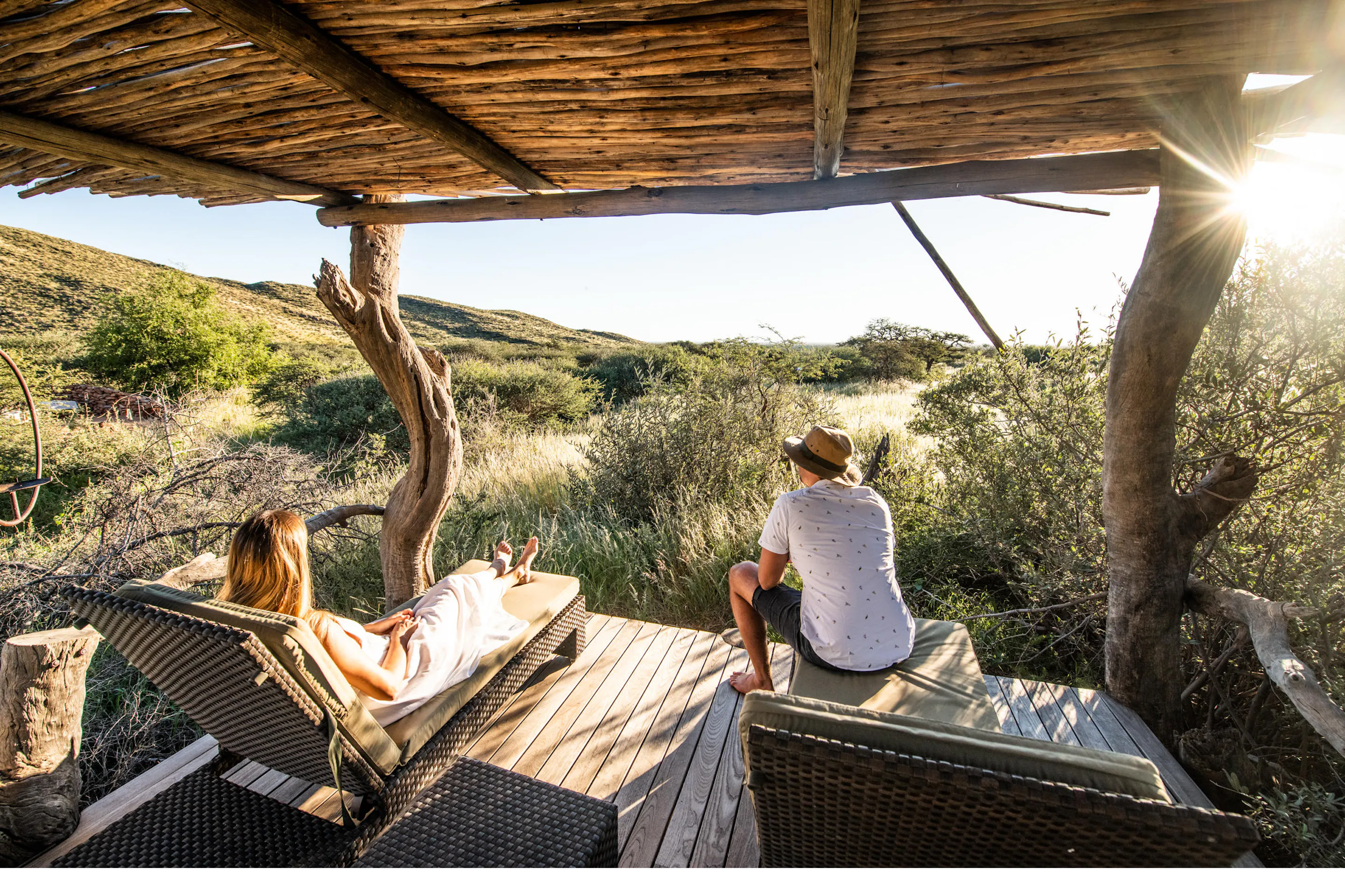 Two guests relax under a thatched shelter, enjoying sundowners as the Kalahari turns gold at sunset, in warm evening light.