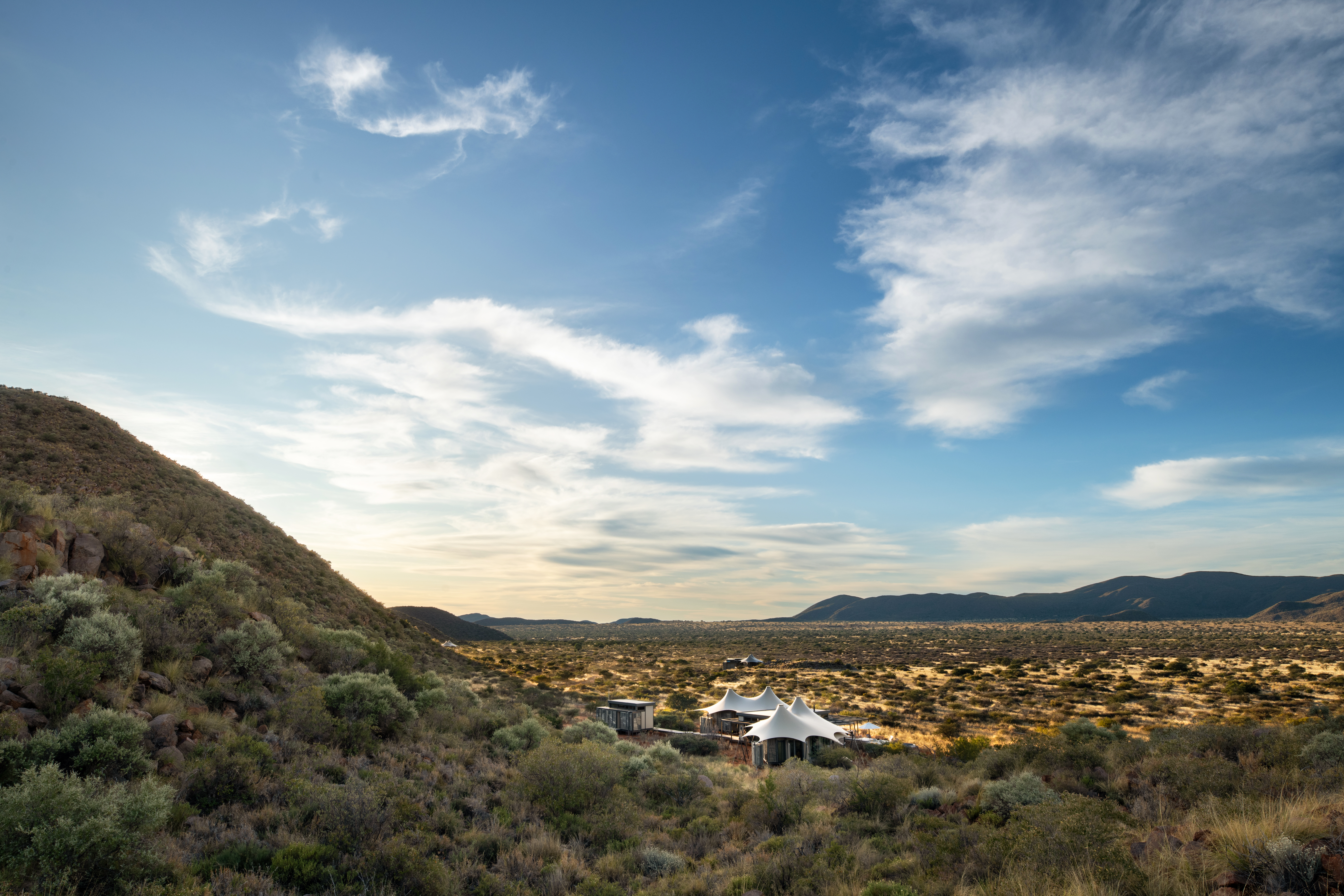 Loapi tents sit in a wide Kalahari valley, low hills and shrubs stretching beneath a bright sky, in warm evening light.
