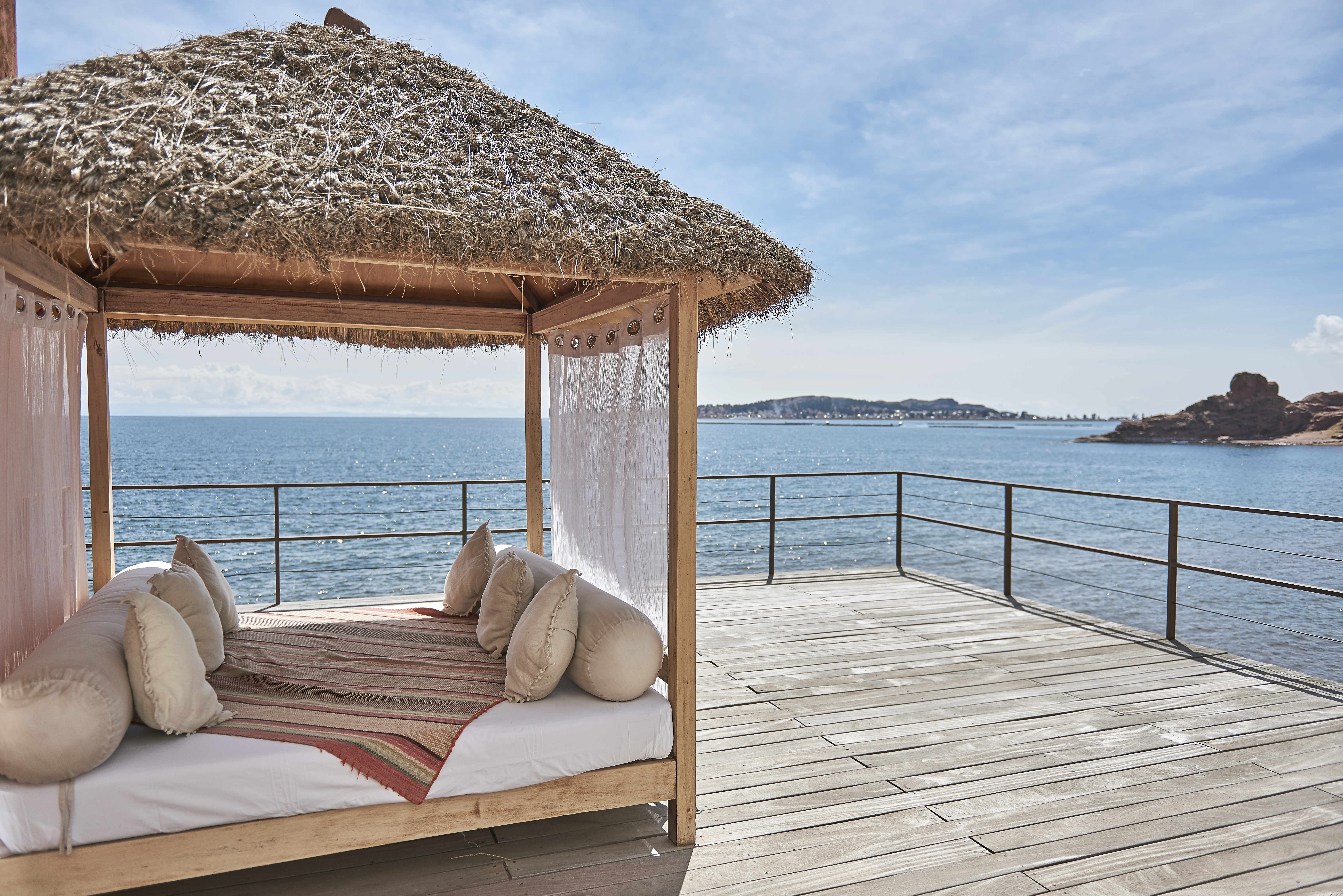 Thatched cabana daybed on a wooden deck overlooks calm Lake Titicaca, with blue water and distant peaks beyond.