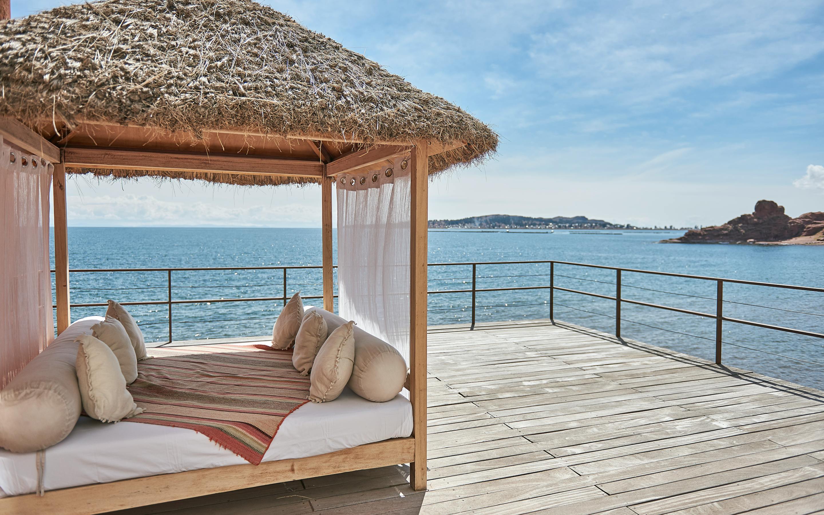 Thatched cabana daybed on a wooden deck overlooks calm Lake Titicaca, with blue water and distant peaks beyond.