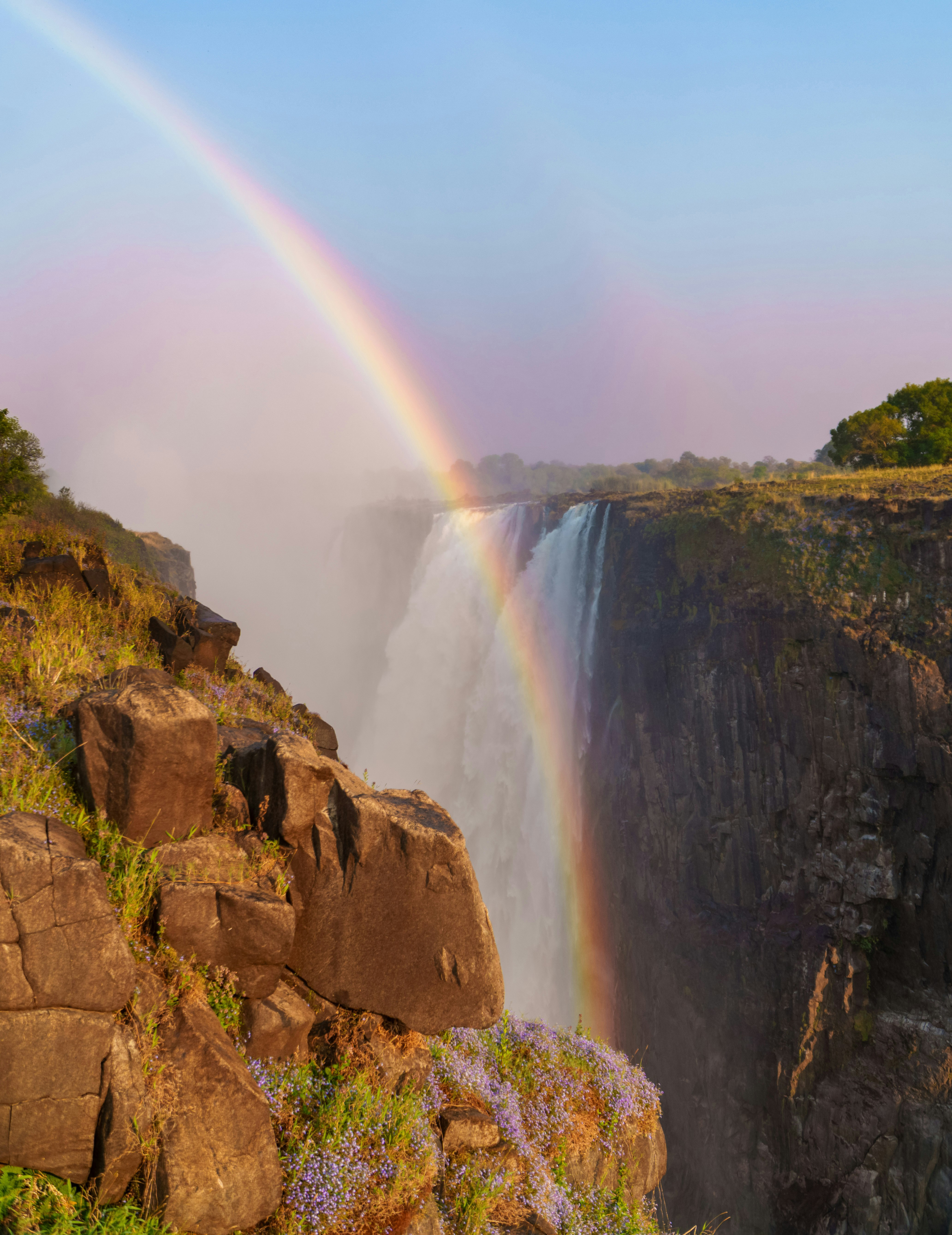 A bright rainbow arcs above Victoria Falls gorge as mist billows up from the chasm beside rugged dark cliffs.