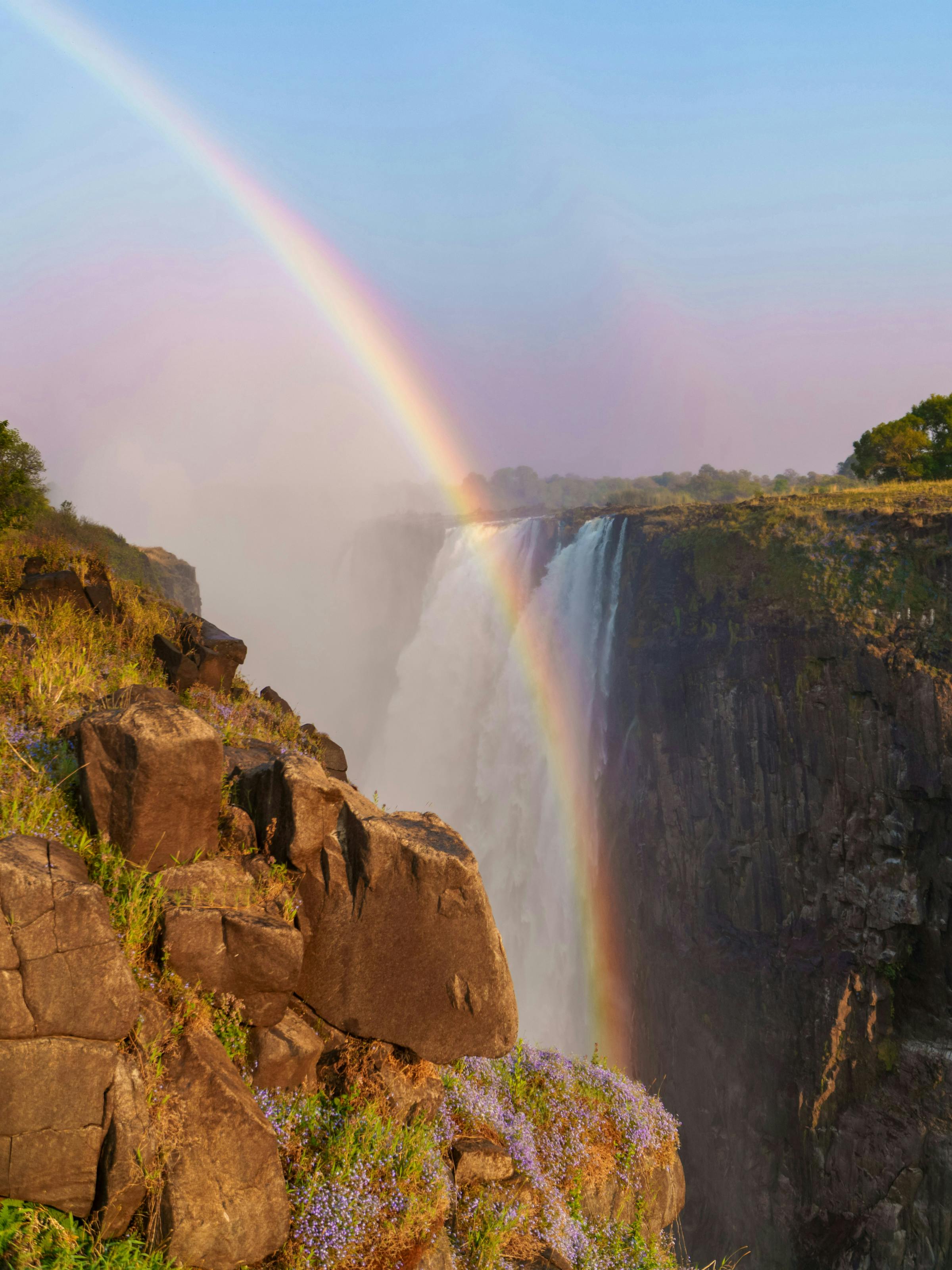 A bright rainbow arcs above Victoria Falls gorge as mist billows up from the chasm beside rugged dark cliffs.