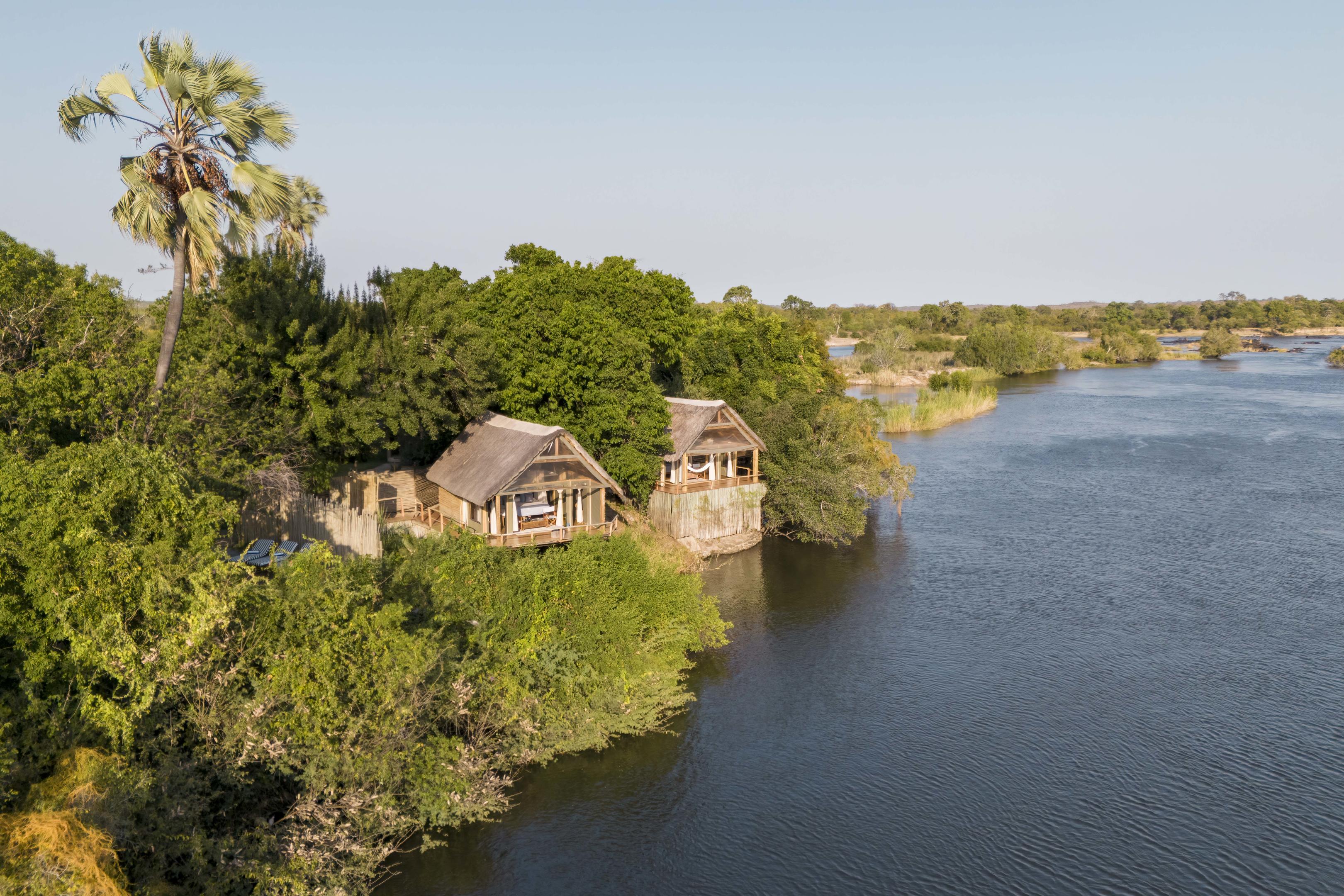 Thatched Sindabezi Lodge chalets sit above the riverbank, shaded by trees with broad Zambezi views beyond.