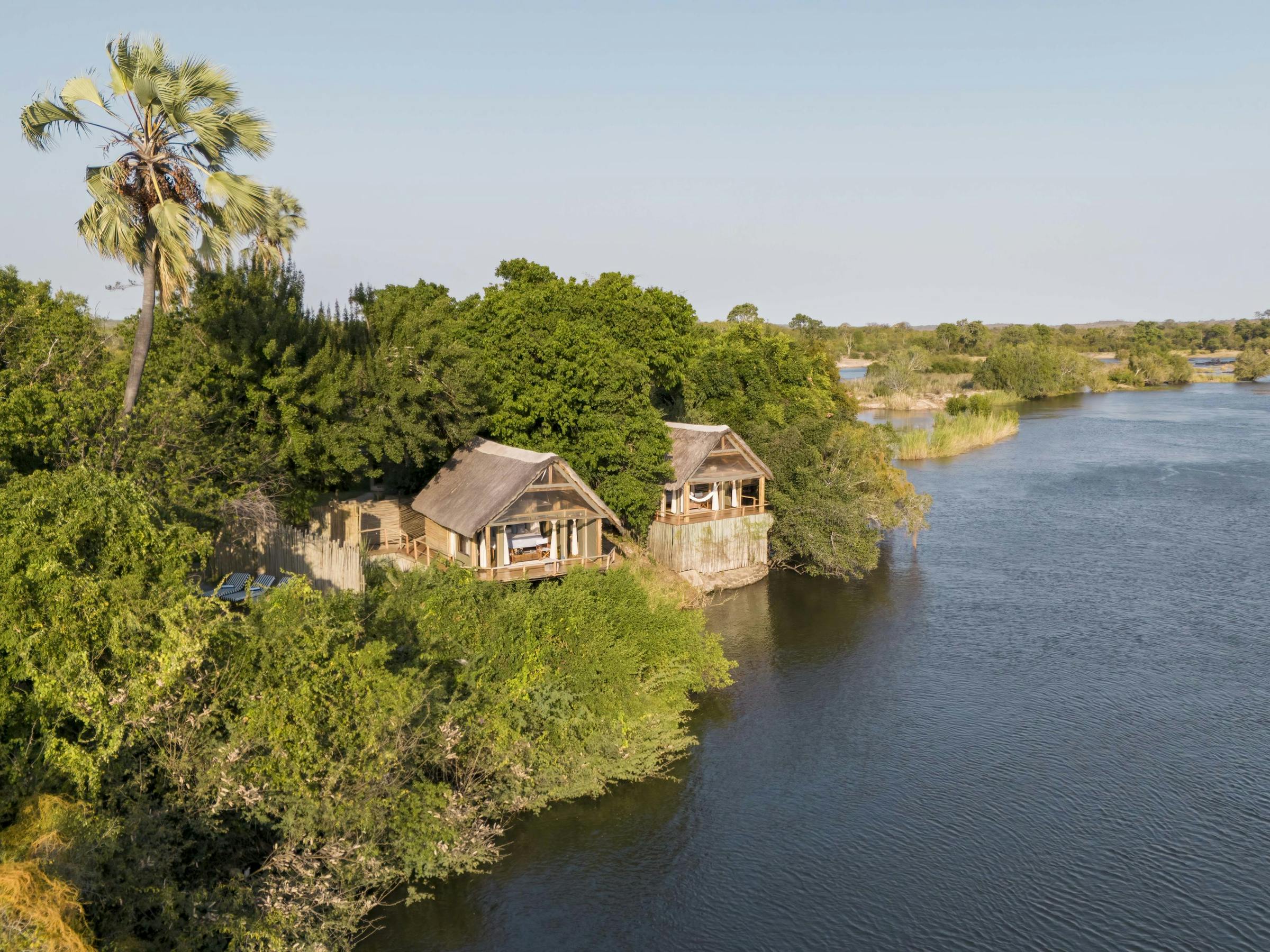 Thatched Sindabezi Lodge chalets sit above the riverbank, shaded by trees with broad Zambezi views beyond.