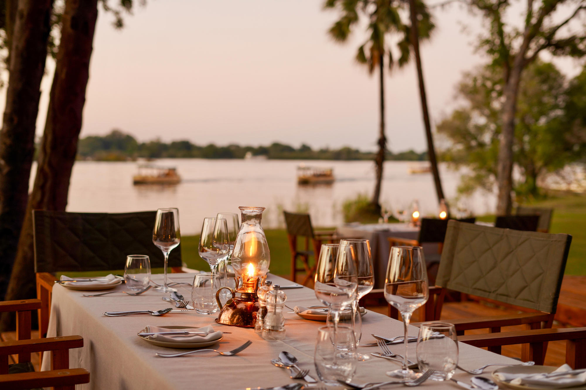 A candlelit table is set for riverside dinner with wine glasses and soft lantern light beside the Zambezi.