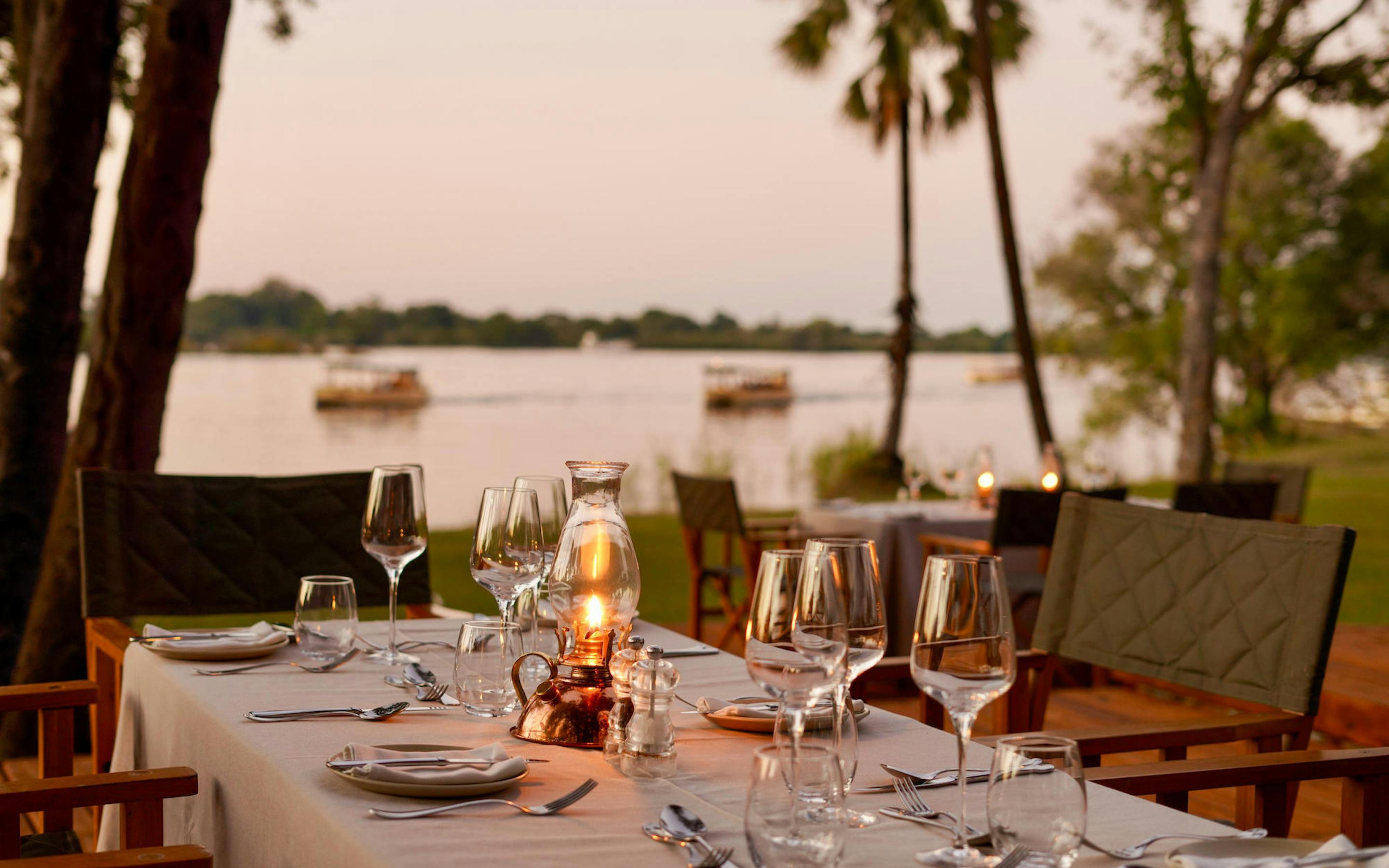 A candlelit table is set for riverside dinner with wine glasses and soft lantern light beside the Zambezi.