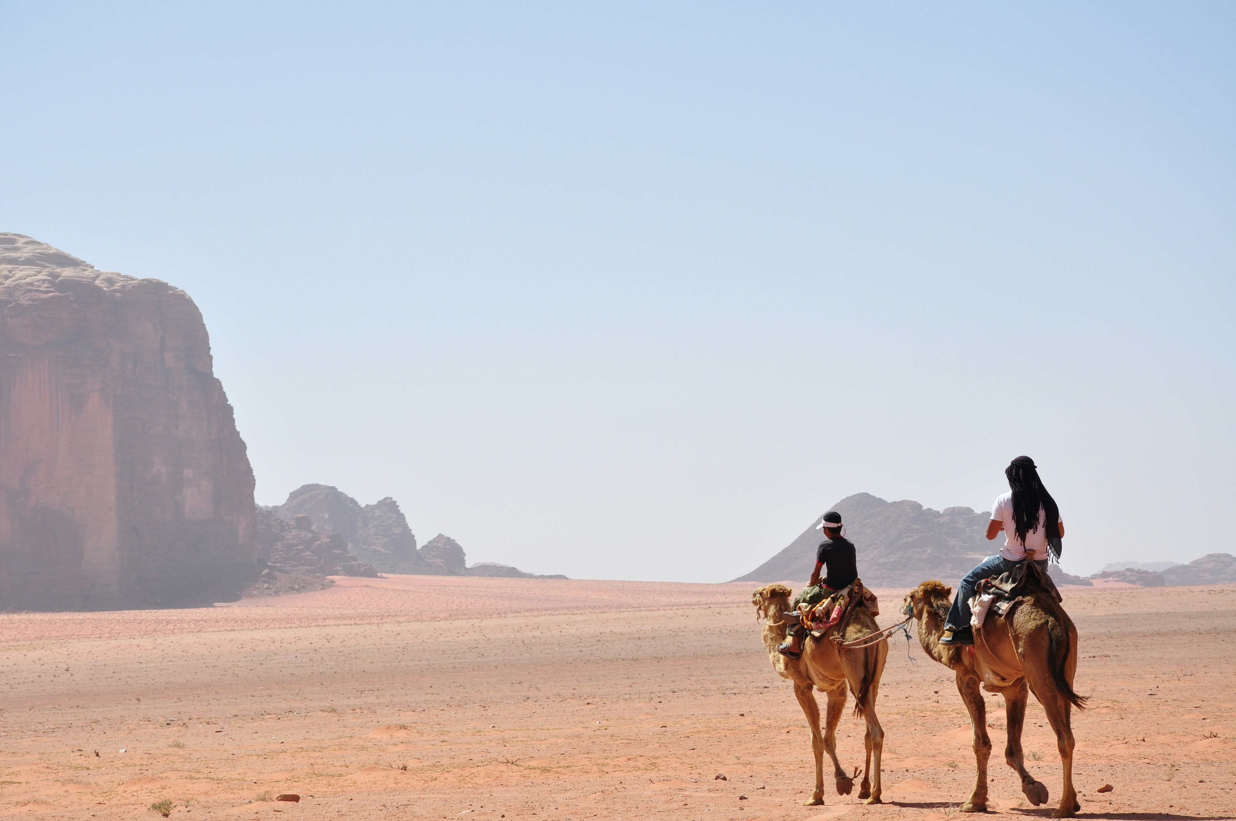 Two camel riders cross Wadi Rum sand at golden hour, long shadows stretching toward distant cliffs and dunes.