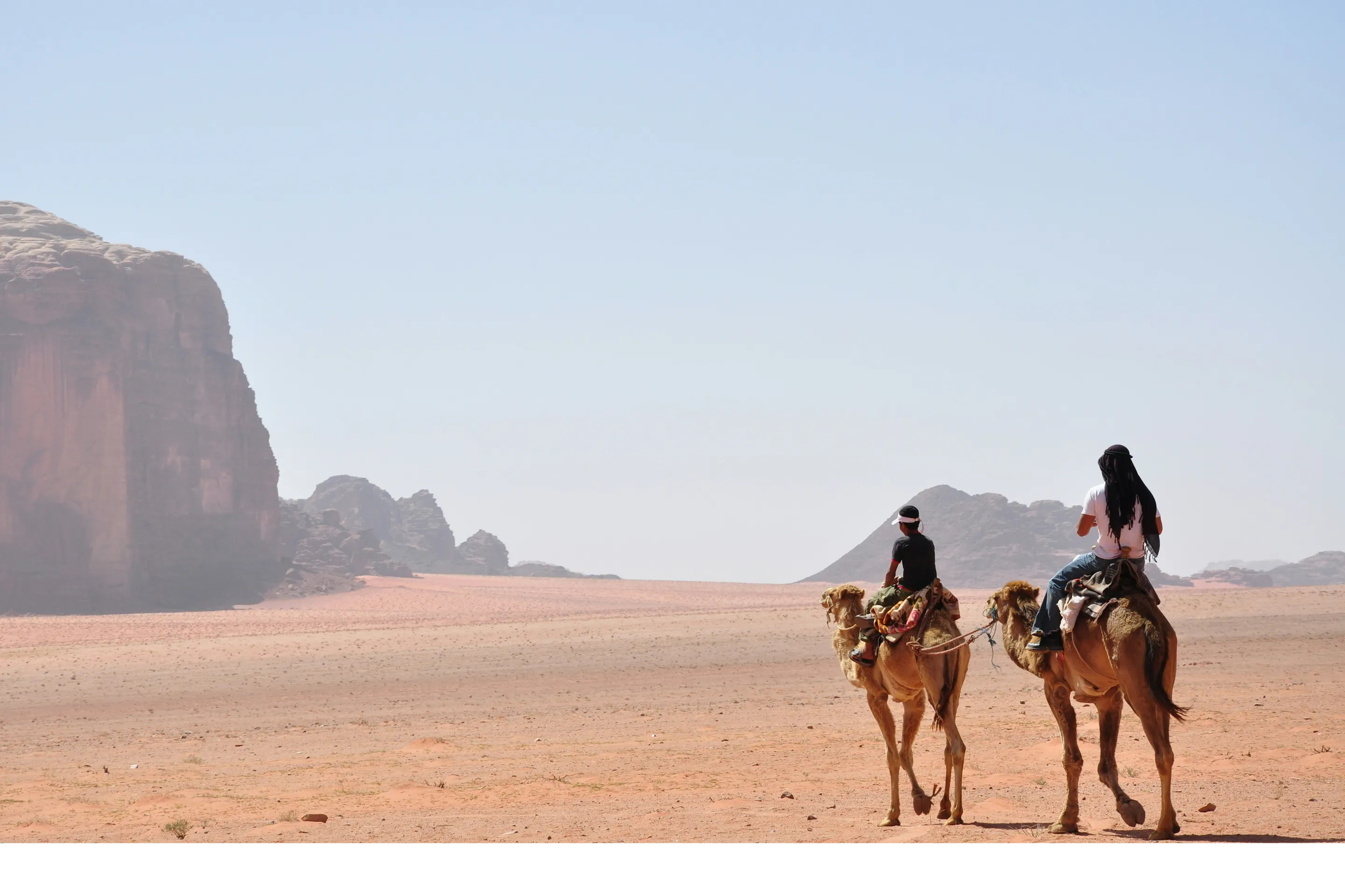 Two camel riders cross Wadi Rum sand at golden hour, long shadows stretching toward distant cliffs and dunes.
