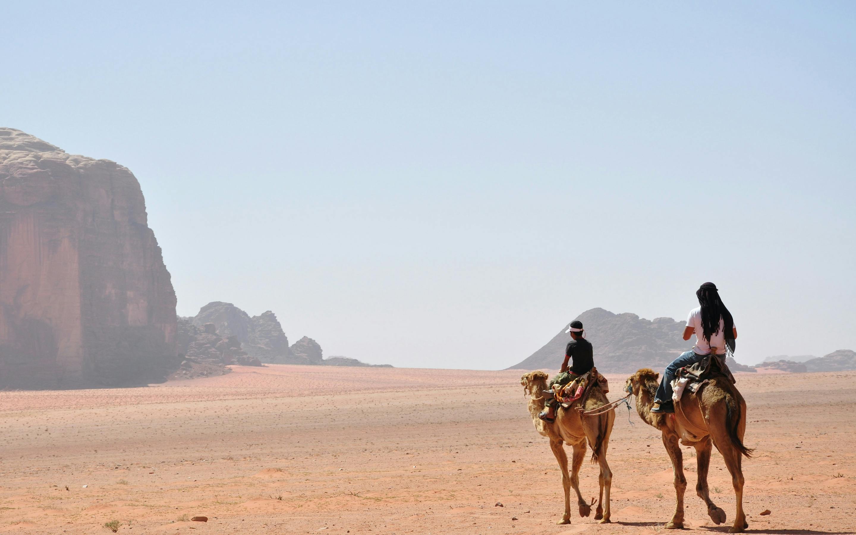 Two camel riders cross Wadi Rum sand at golden hour, long shadows stretching toward distant cliffs and dunes.