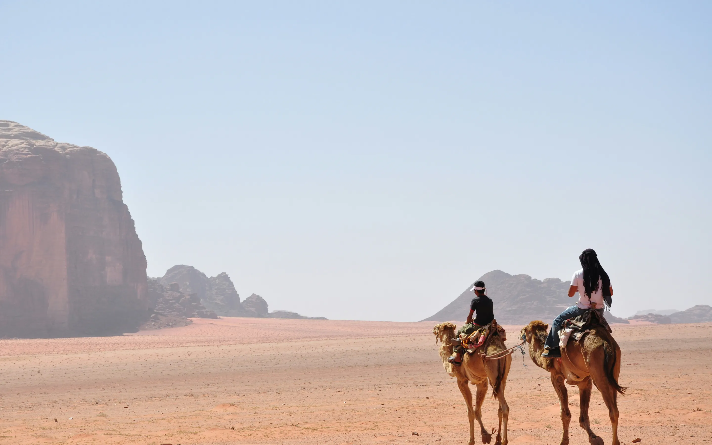 Two camel riders cross Wadi Rum sand at golden hour, long shadows stretching toward distant cliffs and dunes.