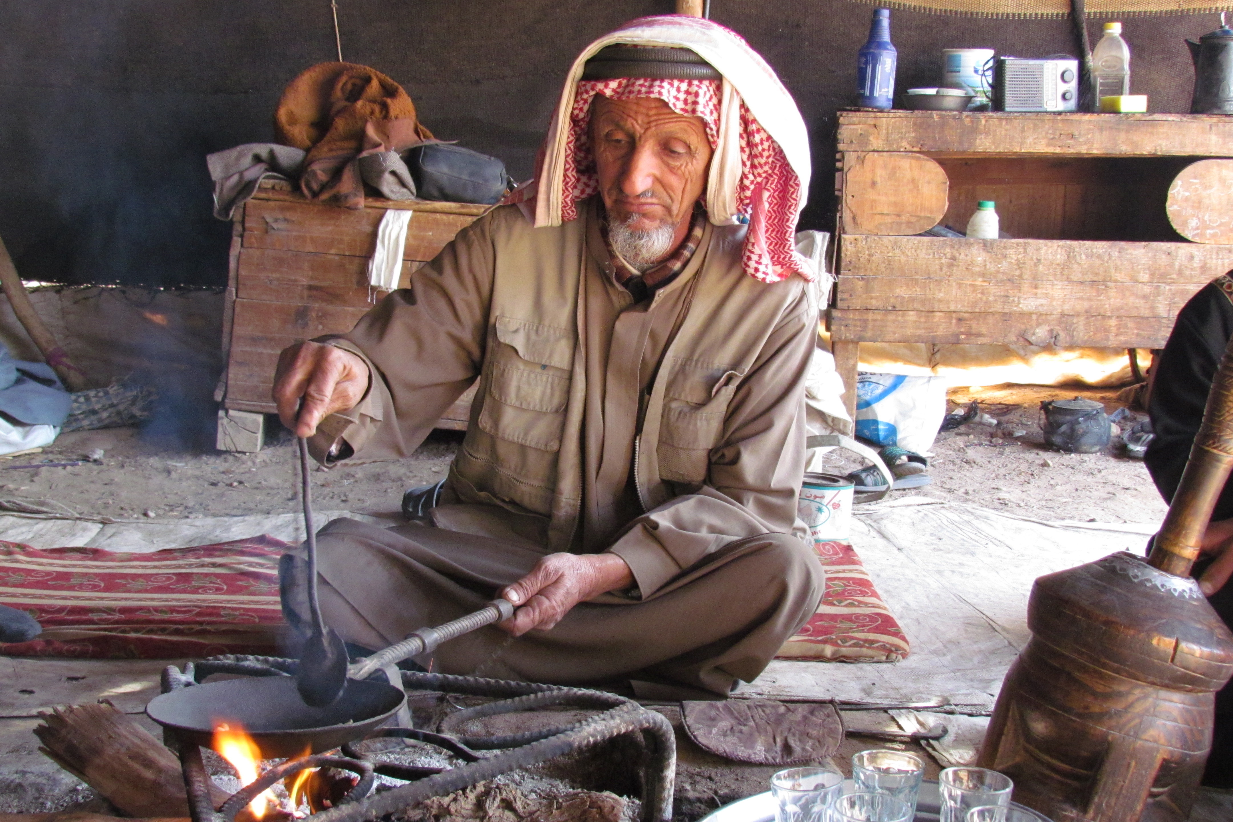A man in a headscarf stirs a shallow pan over embers indoors, with a brass kettle and glasses beside him.