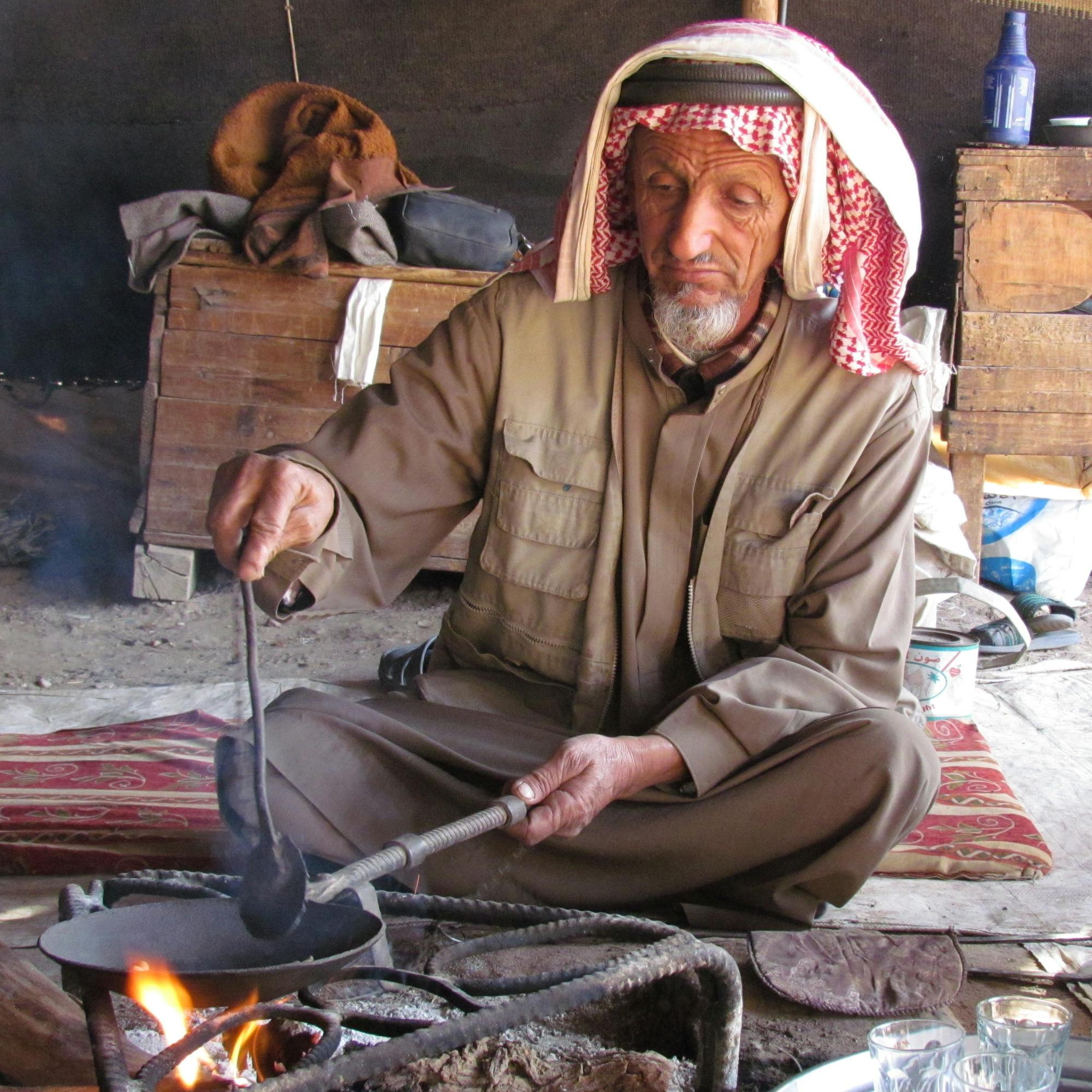 A man in a headscarf stirs a shallow pan over embers indoors, with a brass kettle and glasses beside him.