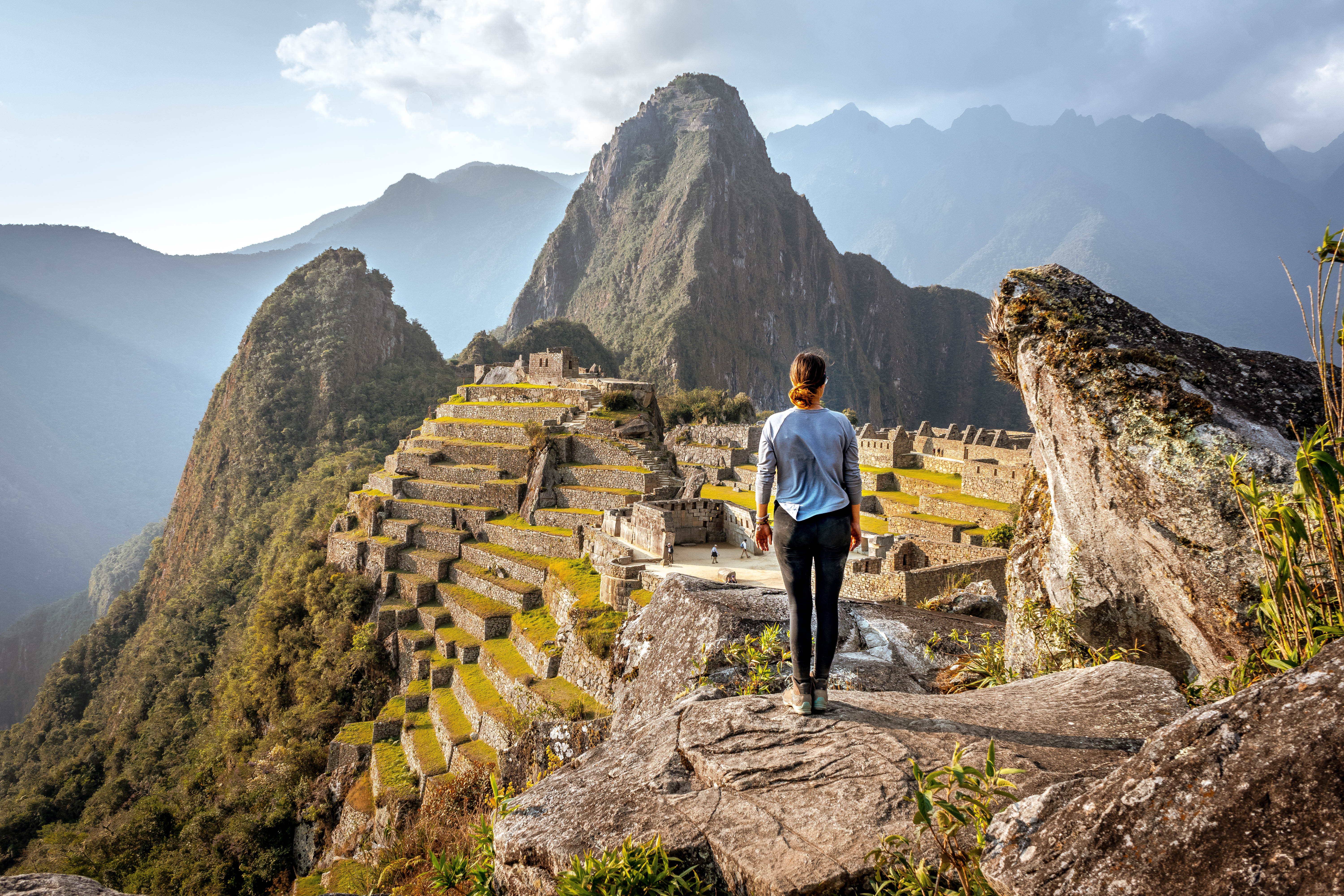 Traveler walks along a stone terrace at Machu Picchu, overlooking stepped ruins and rugged green mountains below.