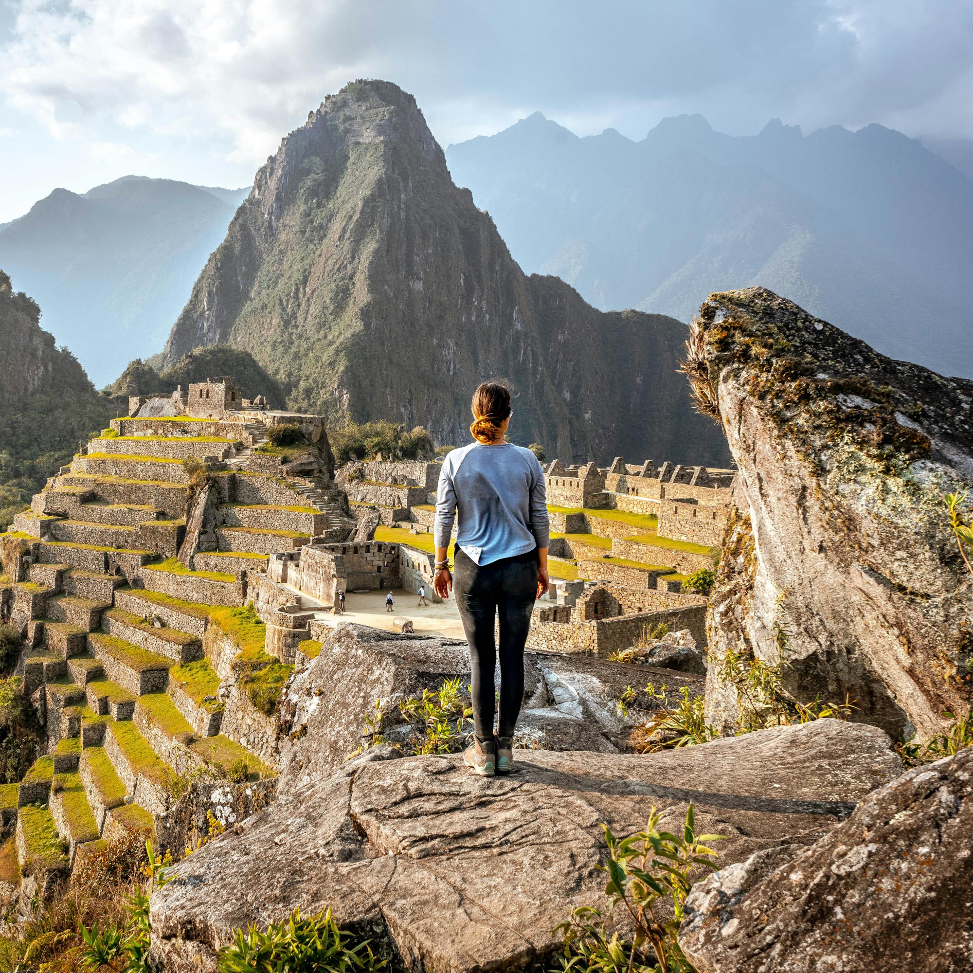 Traveler walks along a stone terrace at Machu Picchu, overlooking stepped ruins and rugged green mountains below.