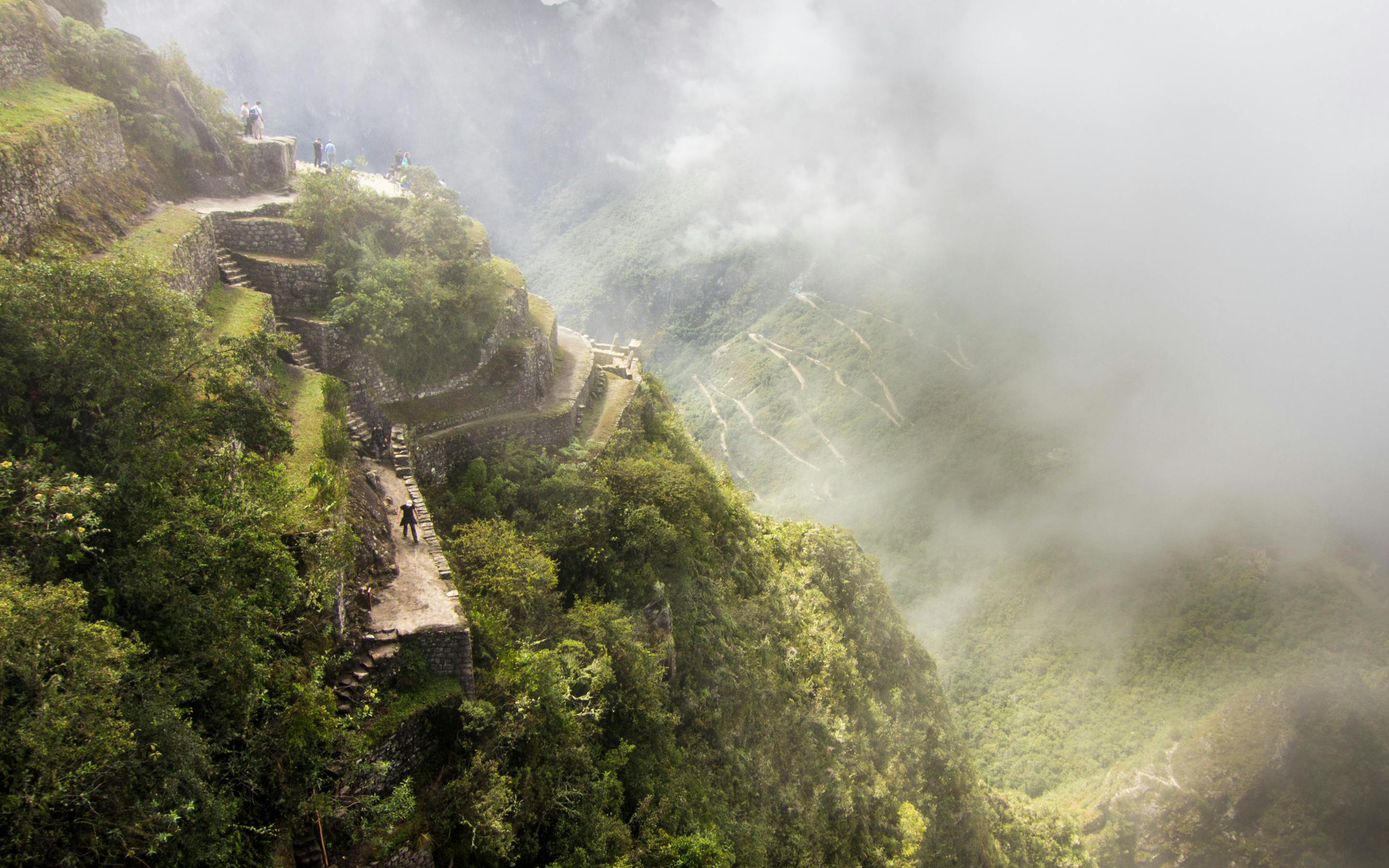 Stone path winds along a misty ridge, with an Inca ruin emerging through sunlit fog above jungle canopy.