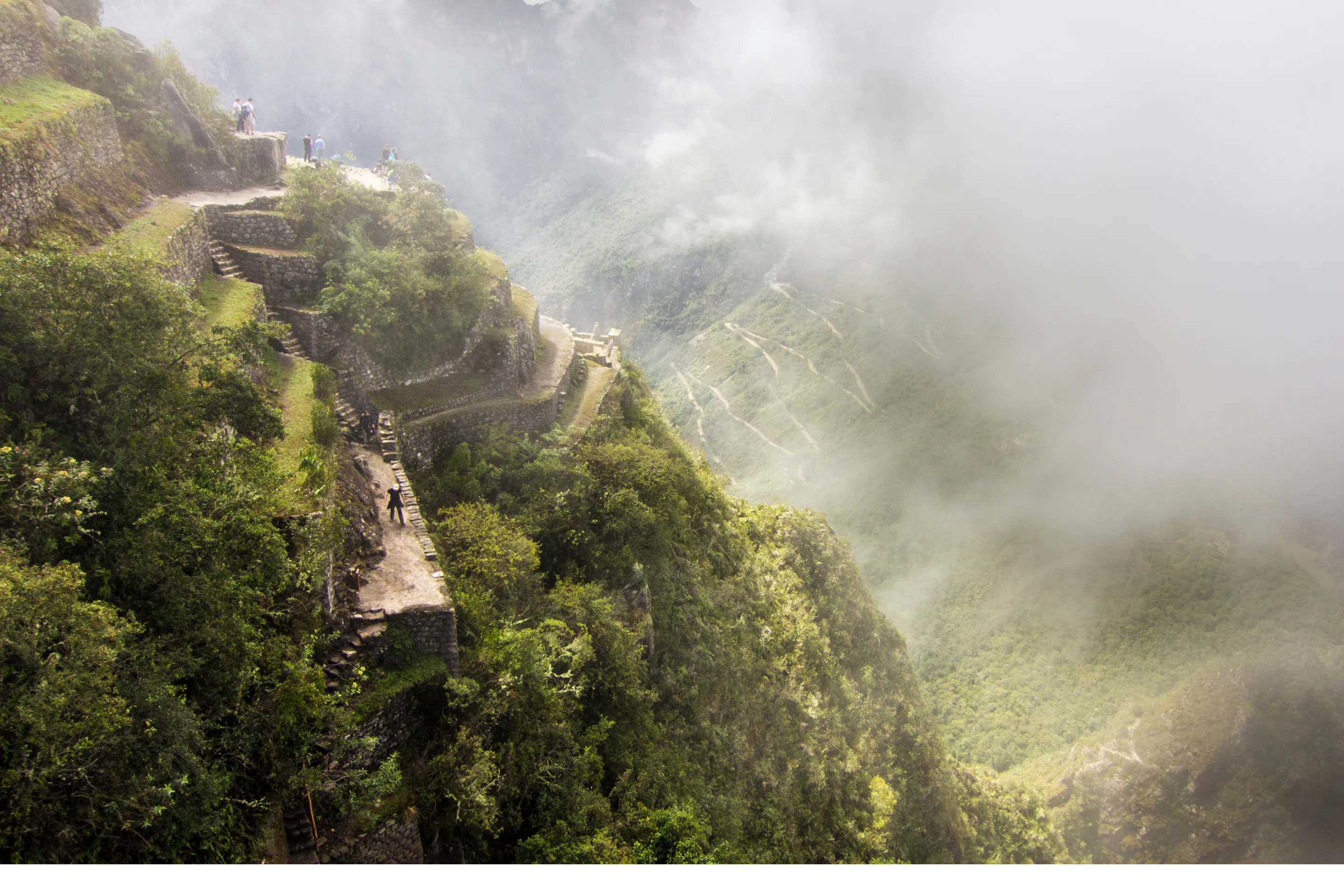 Stone path winds along a misty ridge, with an Inca ruin emerging through sunlit fog above jungle canopy.