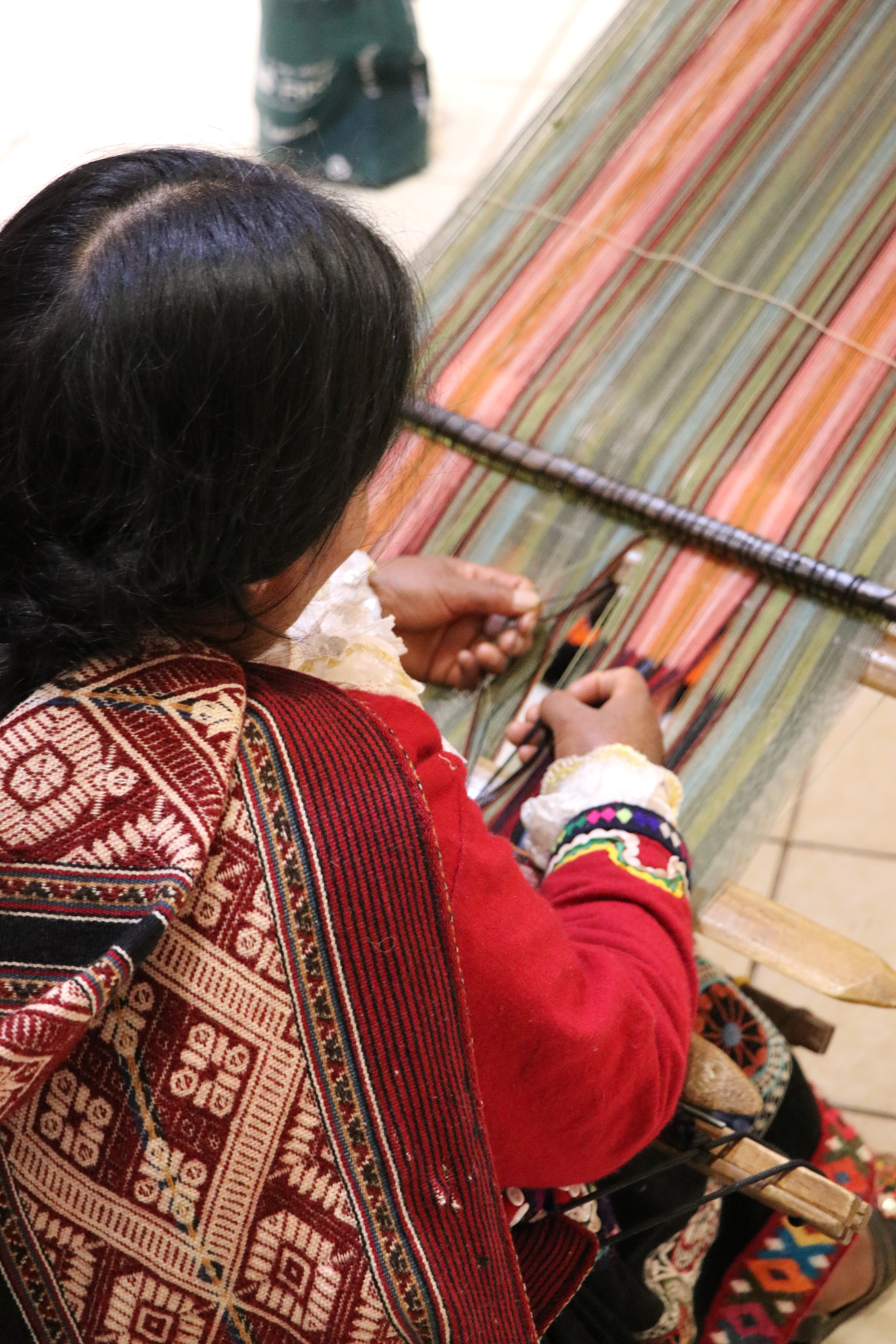 Weaver sits at a loom, hands working colorful threads as patterned textiles drape beside the wooden frame.