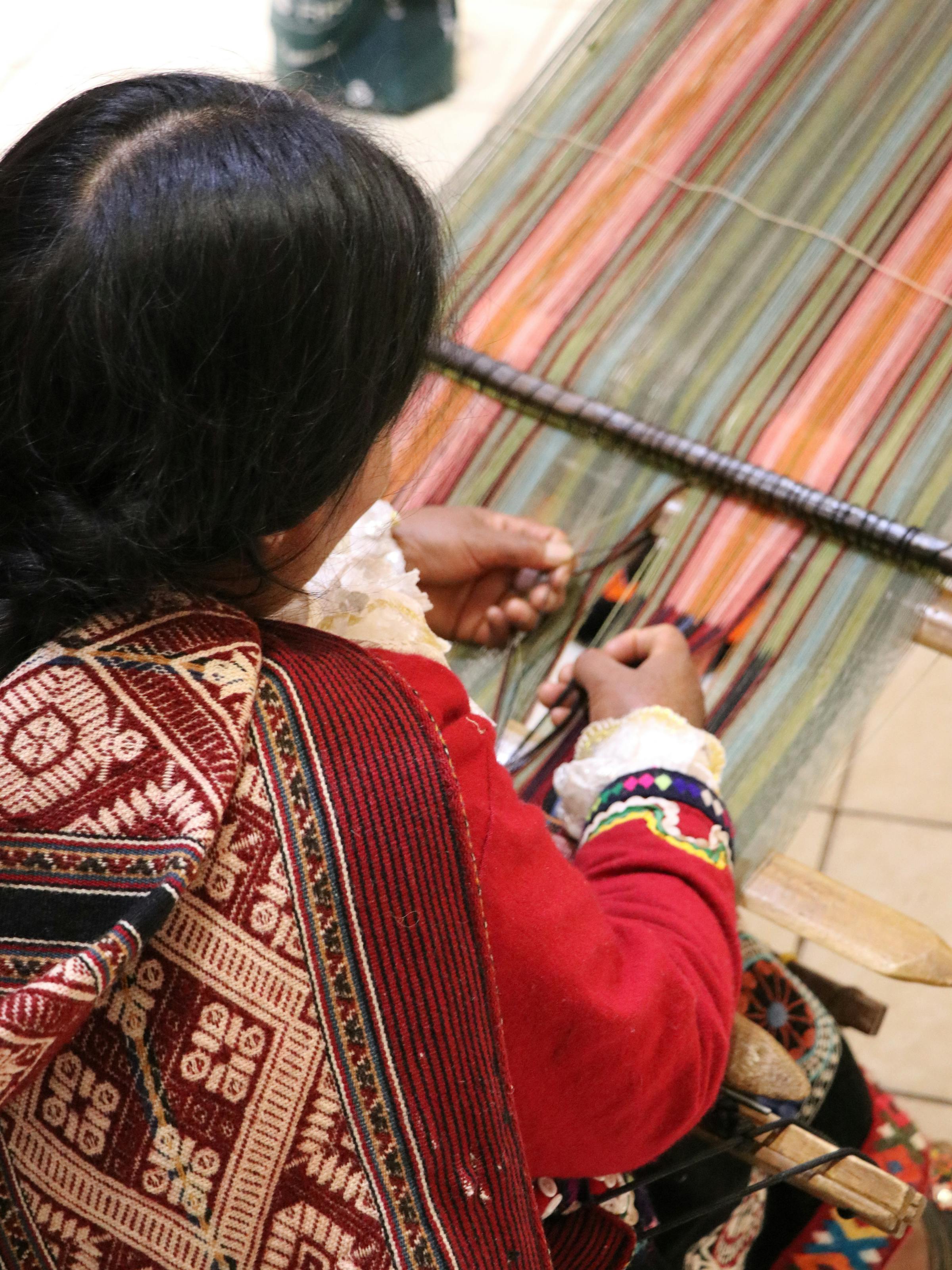 Weaver sits at a loom, hands working colorful threads as patterned textiles drape beside the wooden frame.