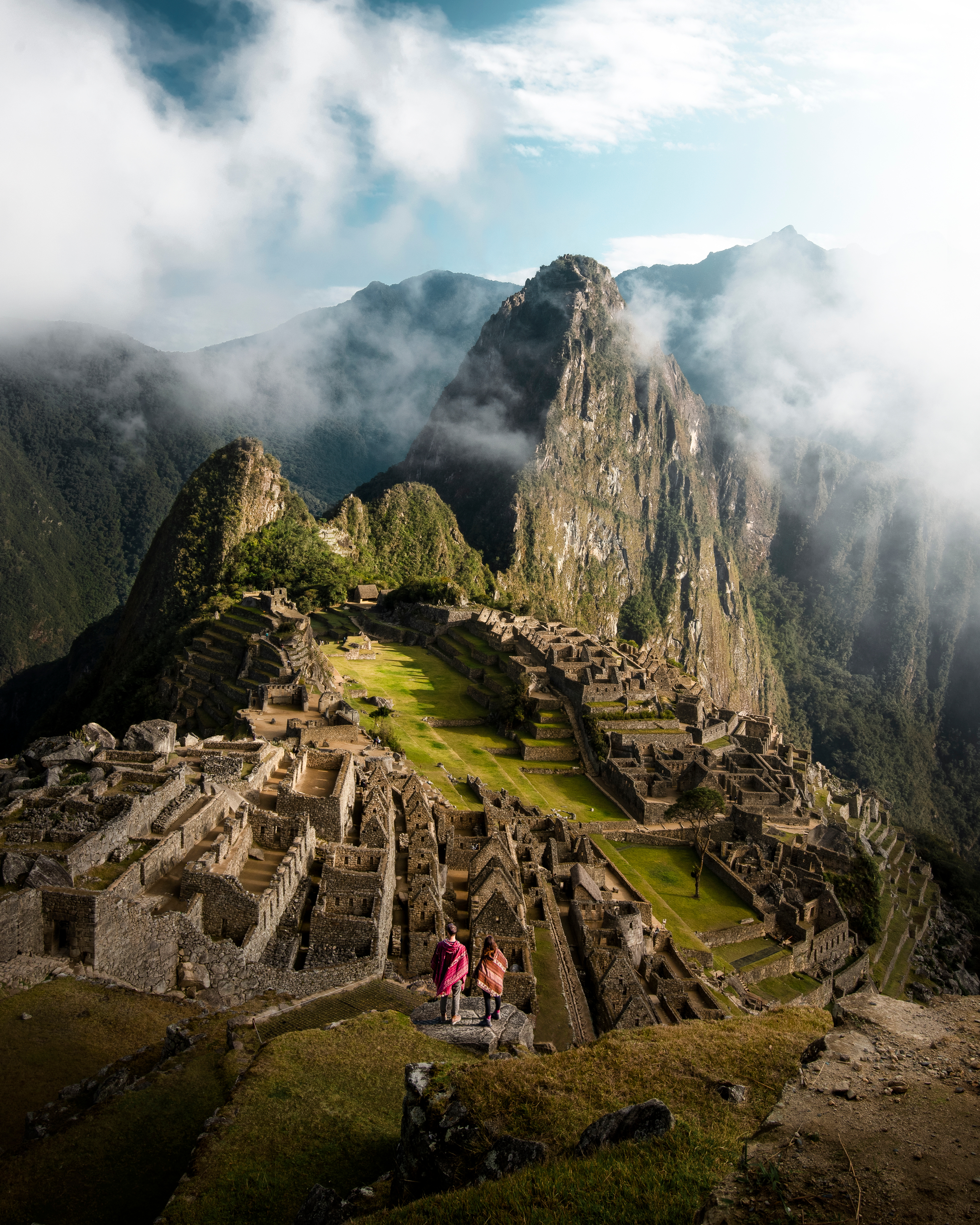 Machu Picchu ruins sit beneath low clouds, stone terraces and green slopes fading into misty mountain air.