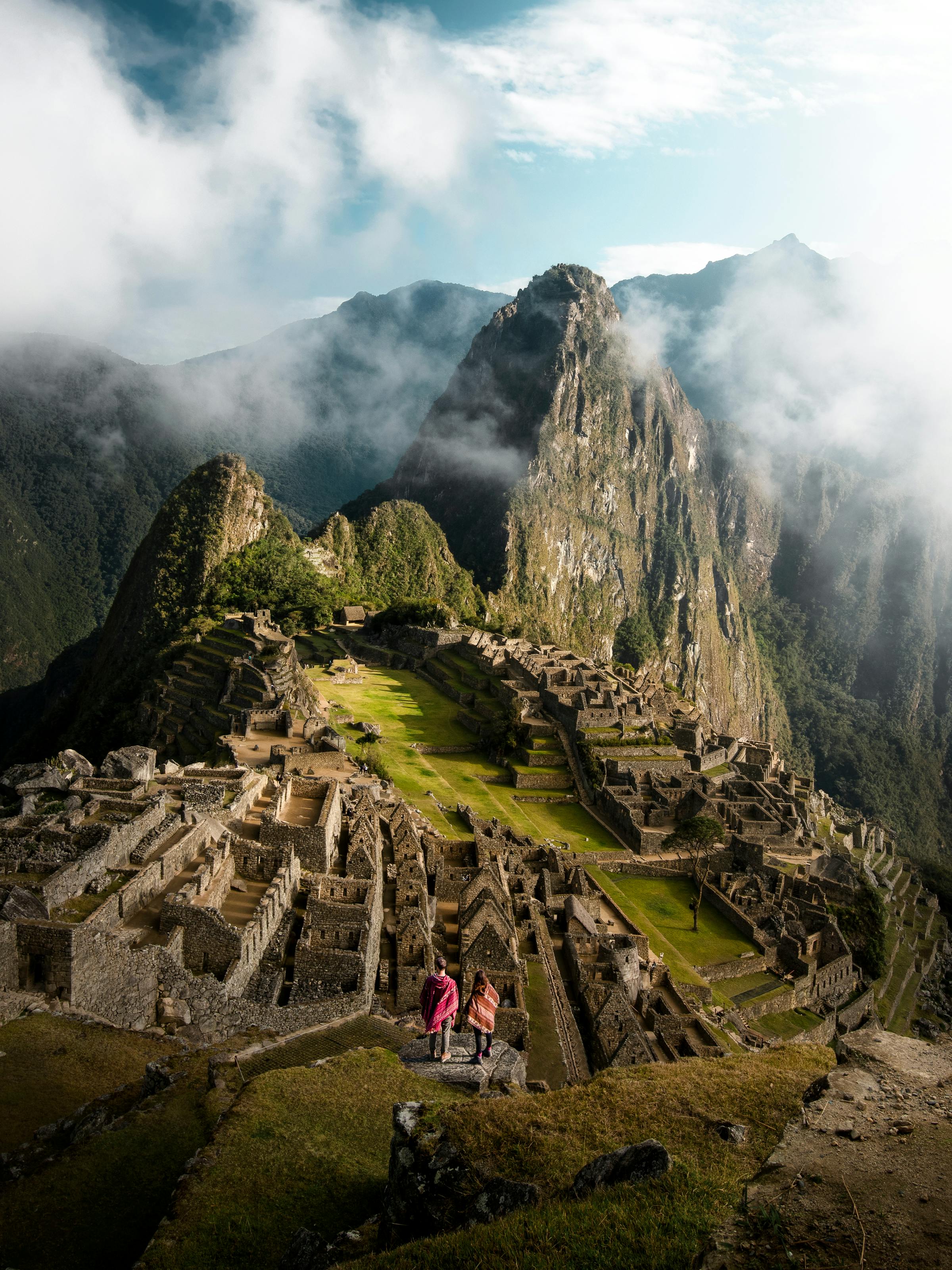 Machu Picchu ruins sit beneath low clouds, stone terraces and green slopes fading into misty mountain air.