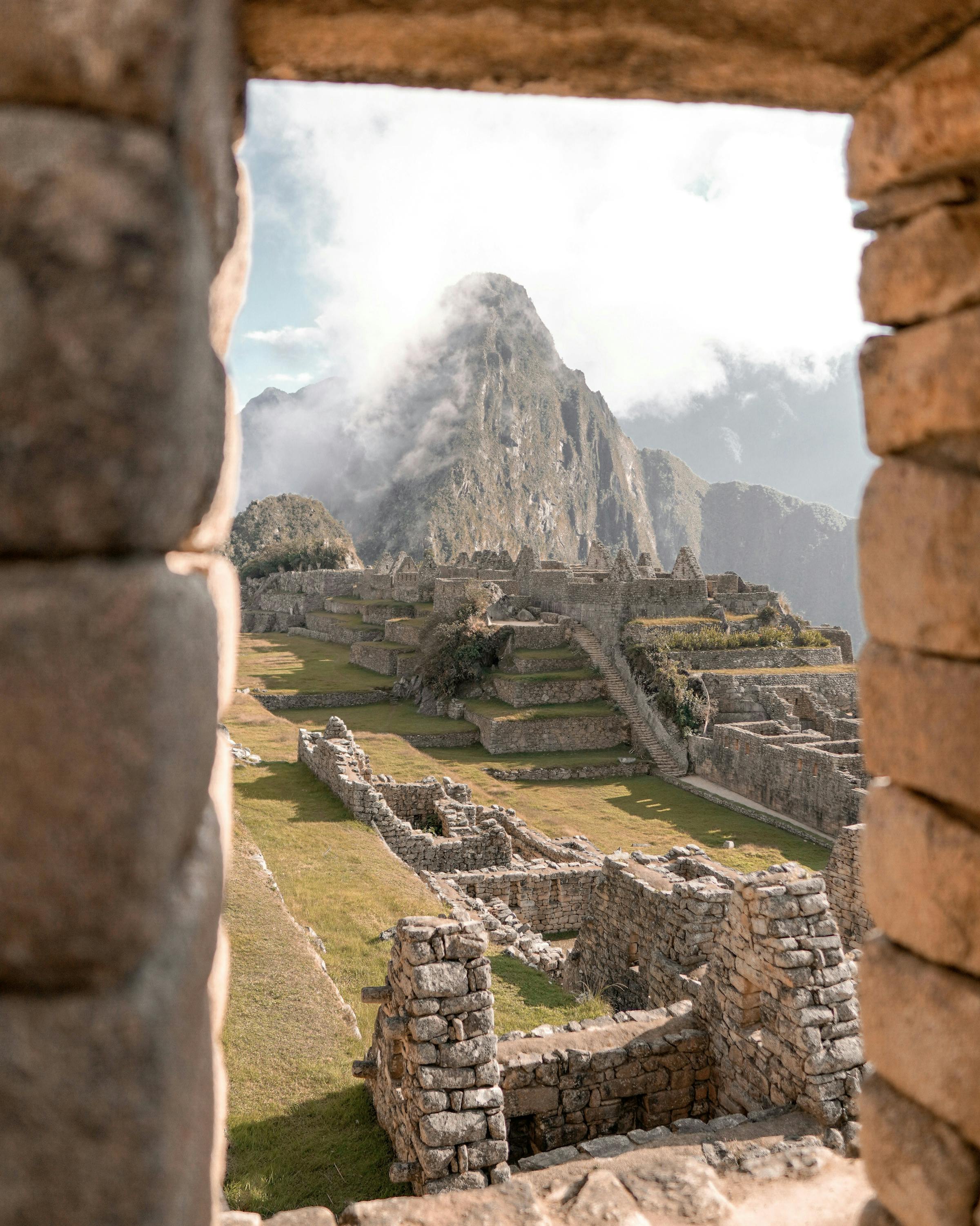 Machu Picchu terraces appear through a stone doorway, framing the ruins and mountains beneath low cloud cover.