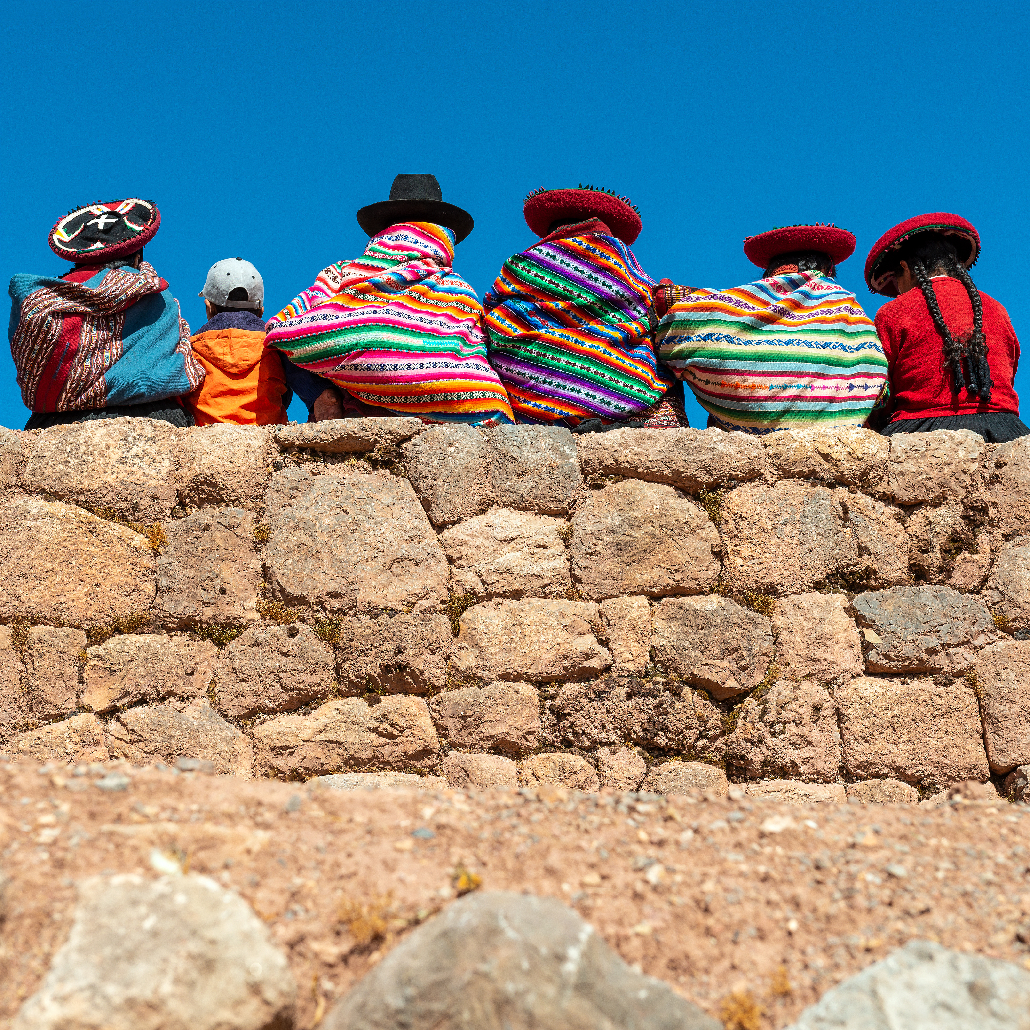 Rows of bright knit hats sit on a stone wall, patterns and pompoms vivid against the clear blue sky.