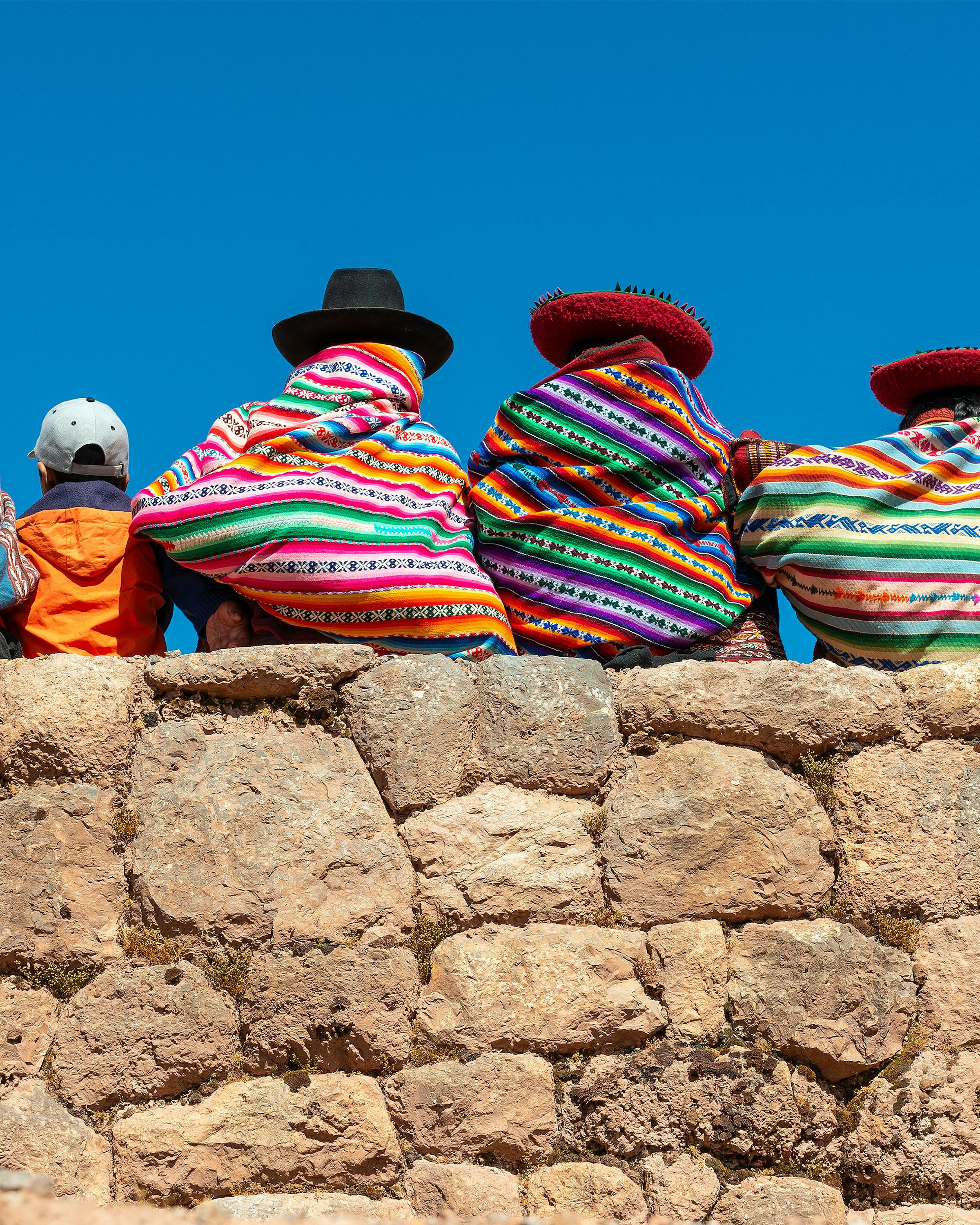 Rows of bright knit hats sit on a stone wall, patterns and pompoms vivid against the clear blue sky.
