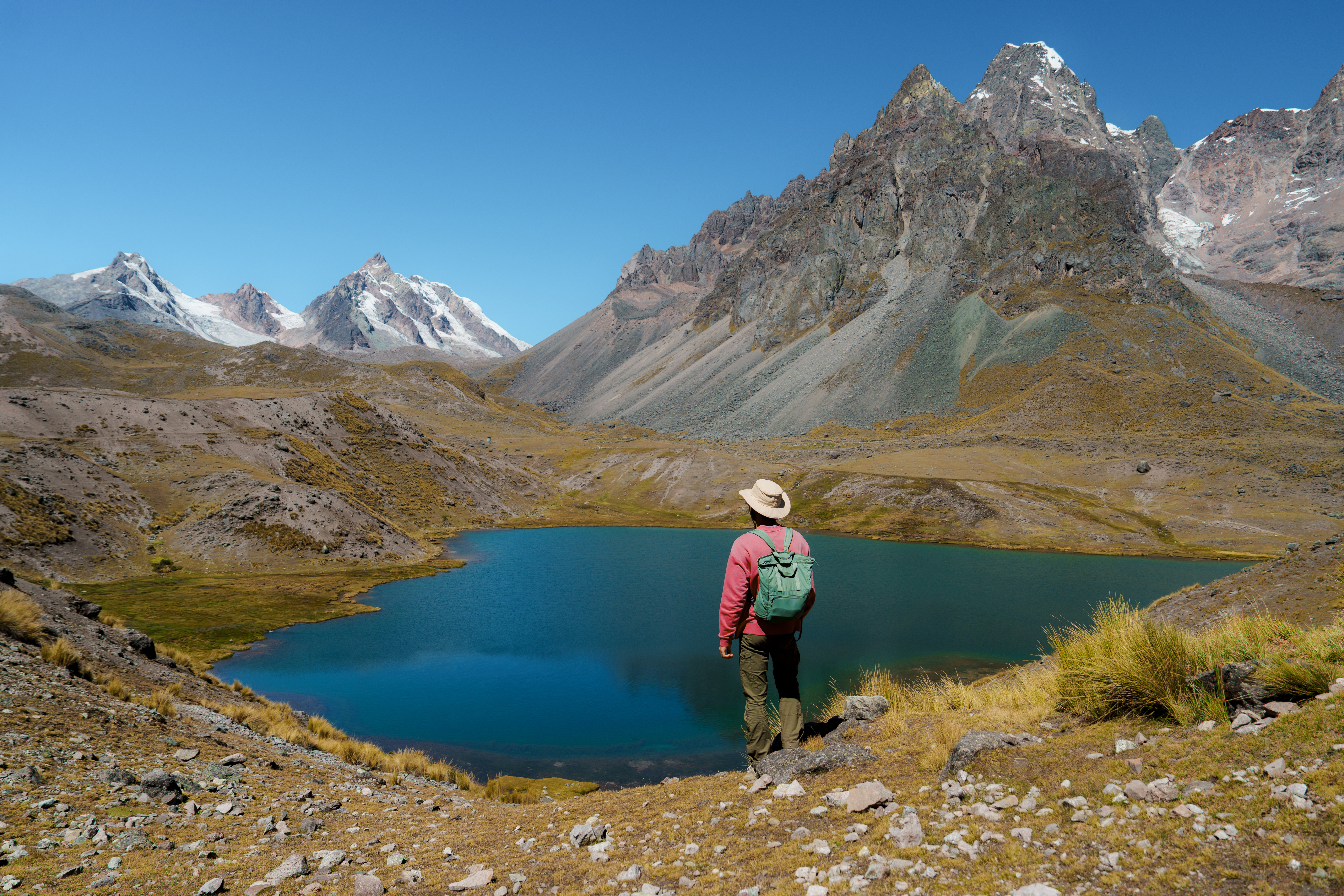 Hiker stands beside a turquoise mountain lagoon, snowcapped peaks and rocky slopes towering above the water.
