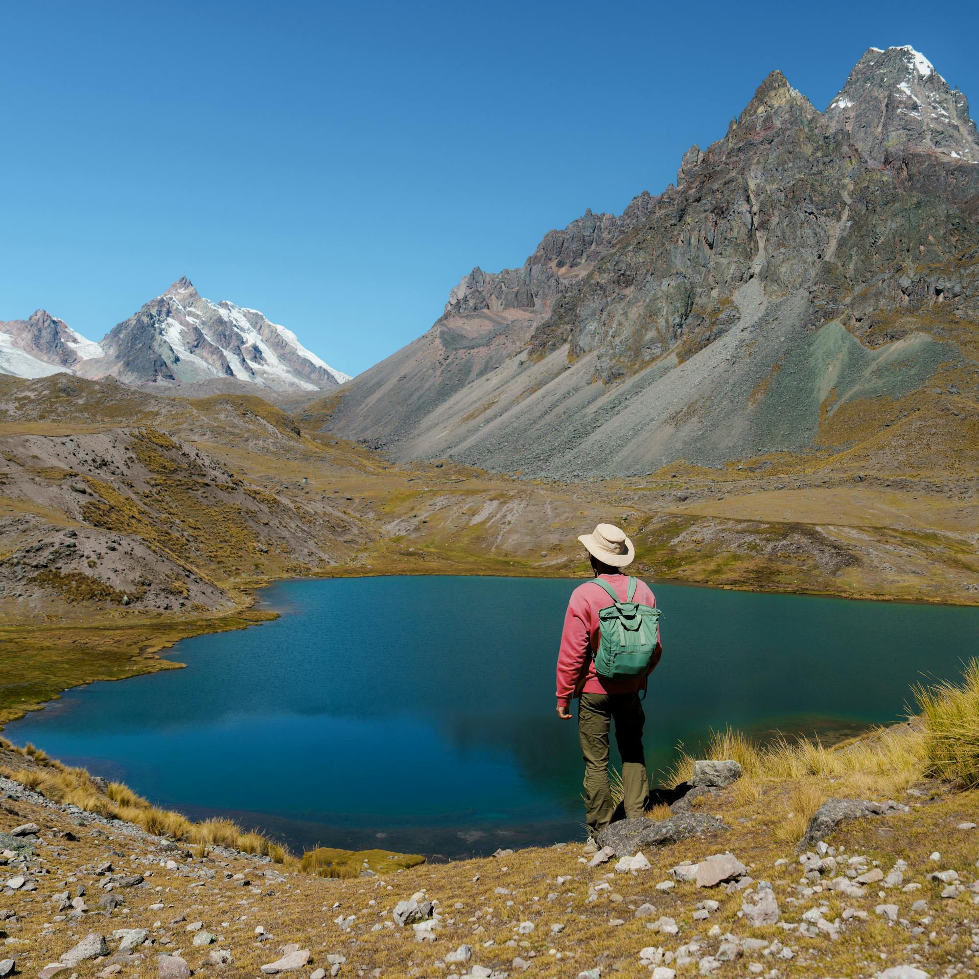 Hiker stands beside a turquoise mountain lagoon, snowcapped peaks and rocky slopes towering above the water.