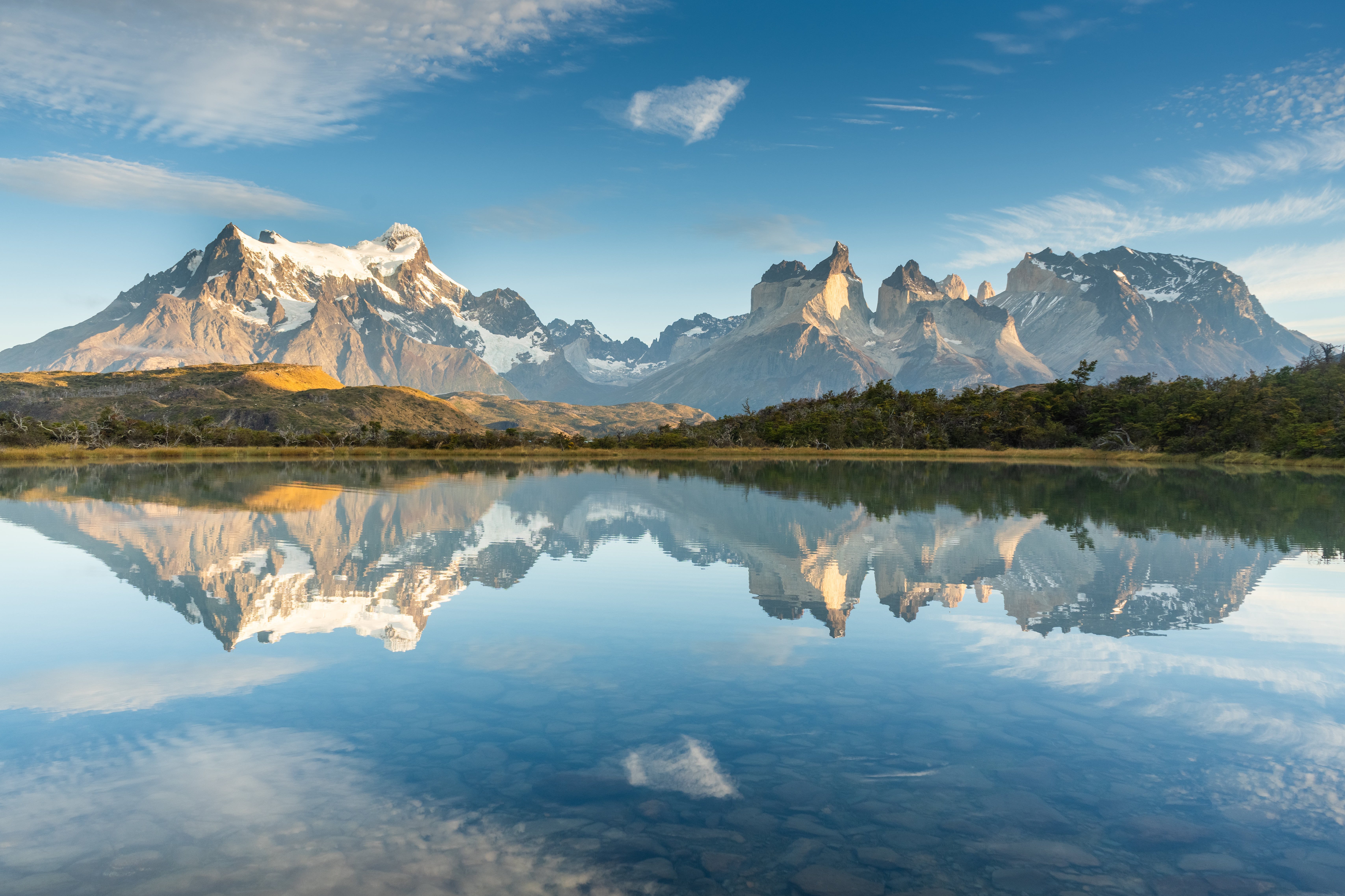 Snow-capped mountain peaks rising above a calm lake, with their reflections mirrored in the clear water under a bright blue sky with scattered clouds.