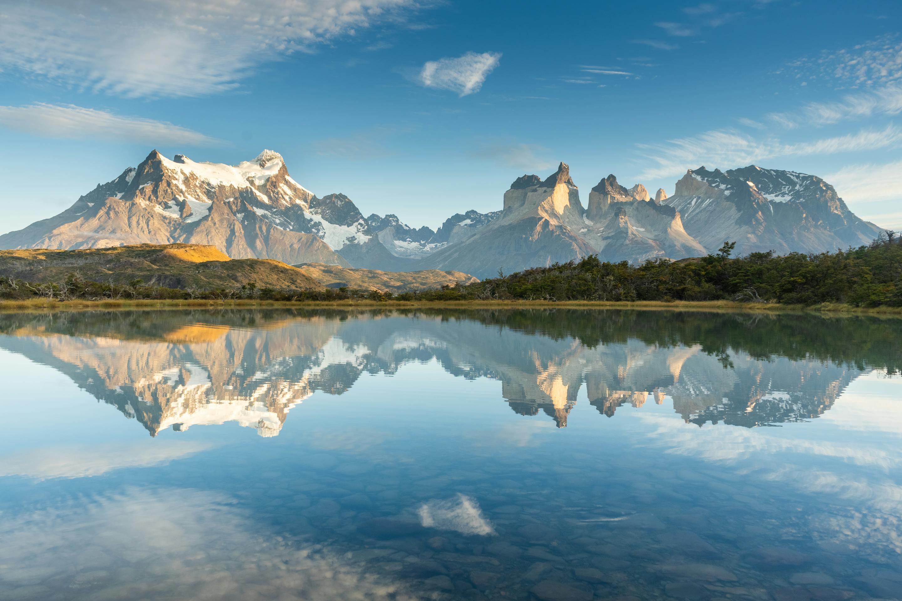 Snow-capped mountain peaks rising above a calm lake, with their reflections mirrored in the clear water under a bright blue sky with scattered clouds.