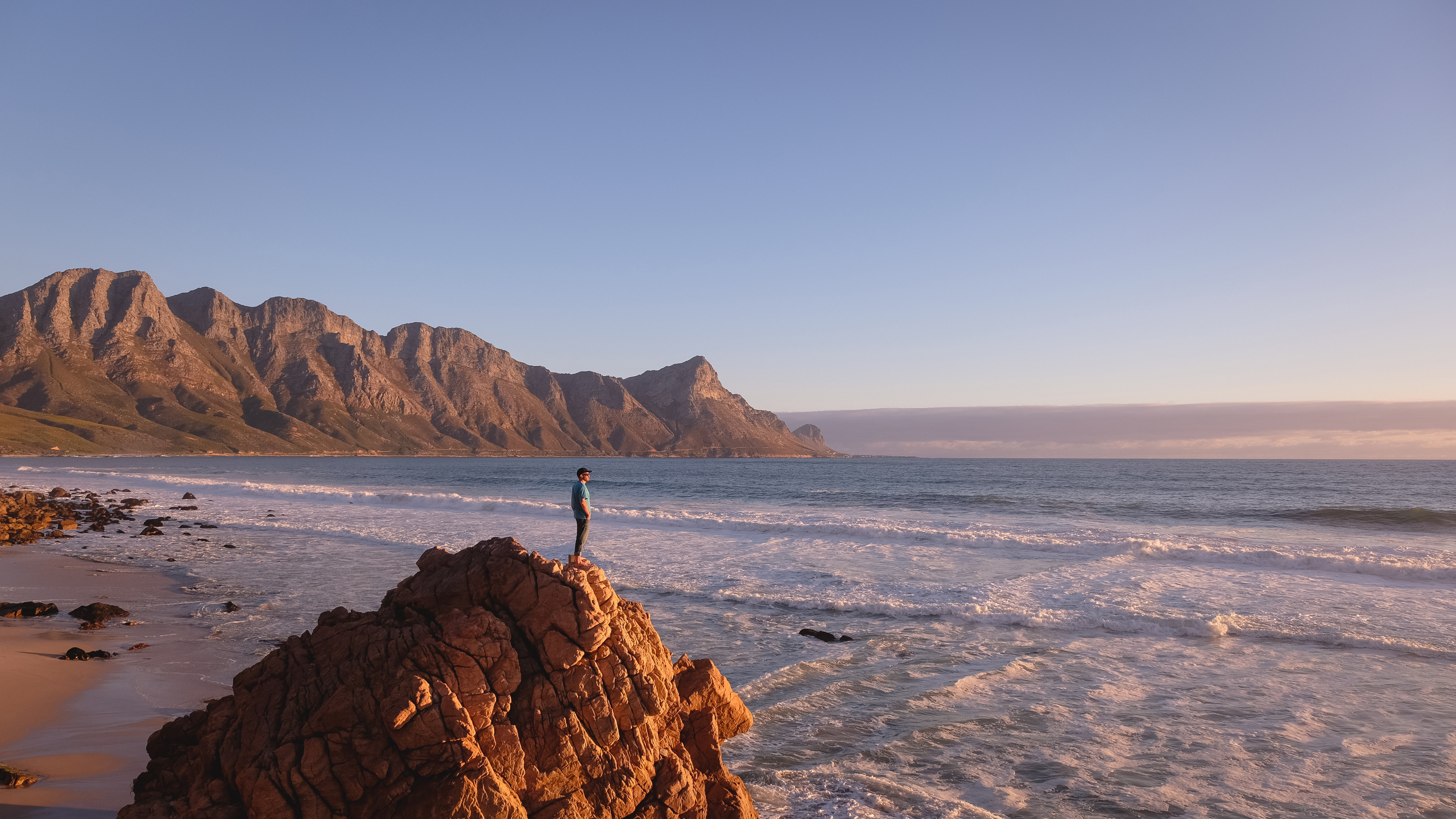 A lone traveler stands on coastal rocks above rolling surf, with a long sandy bay and mountains under pastel sky.